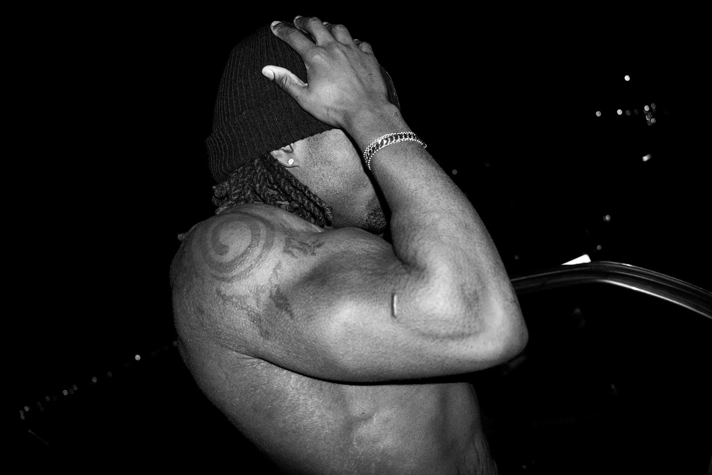 A shirtless man with tattoos on his arm, wearing a beanie and bracelet, covering his face with his hand against a dark background with small lights.