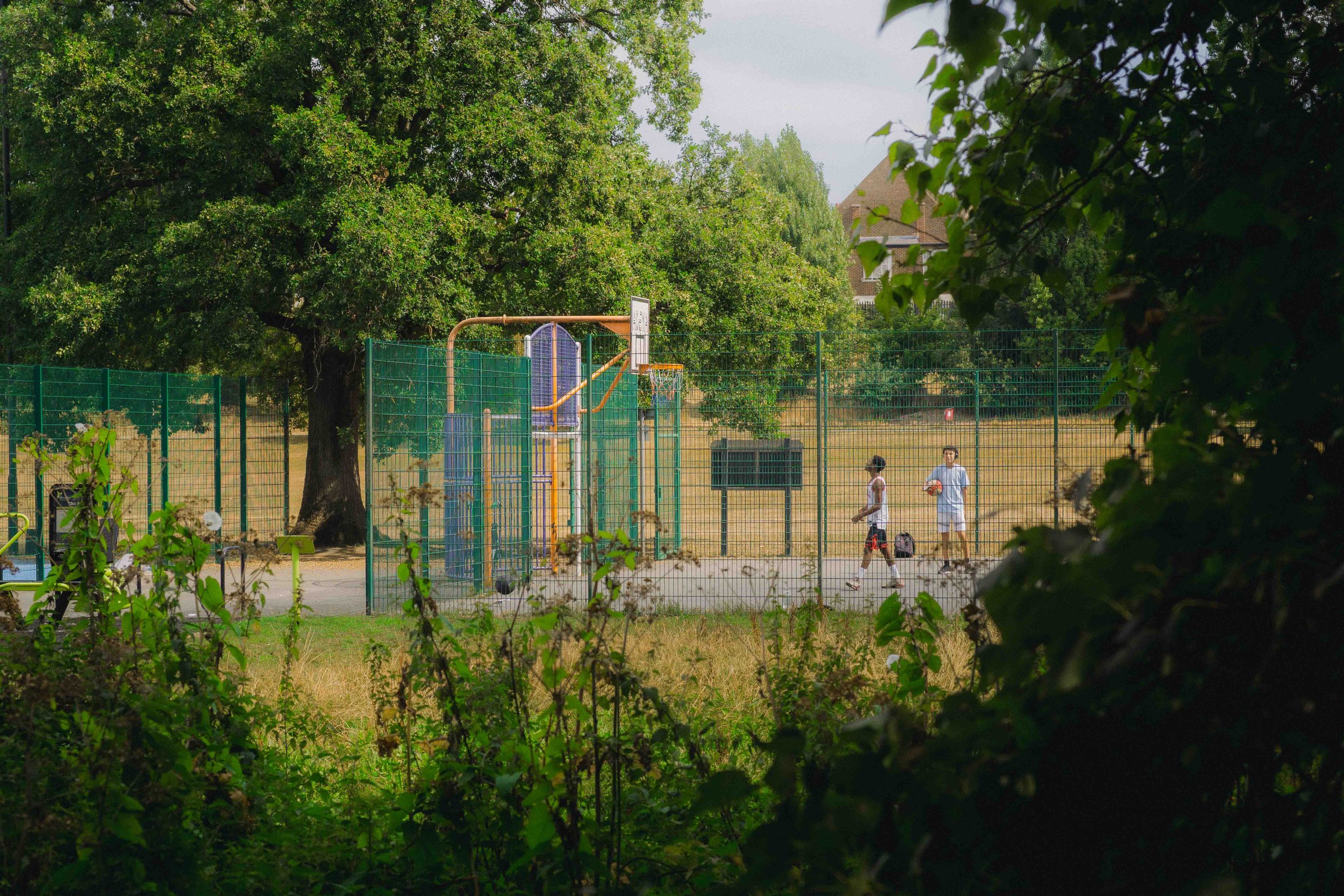 People playing basketball on an outdoor court, surrounded by trees and a green fence.