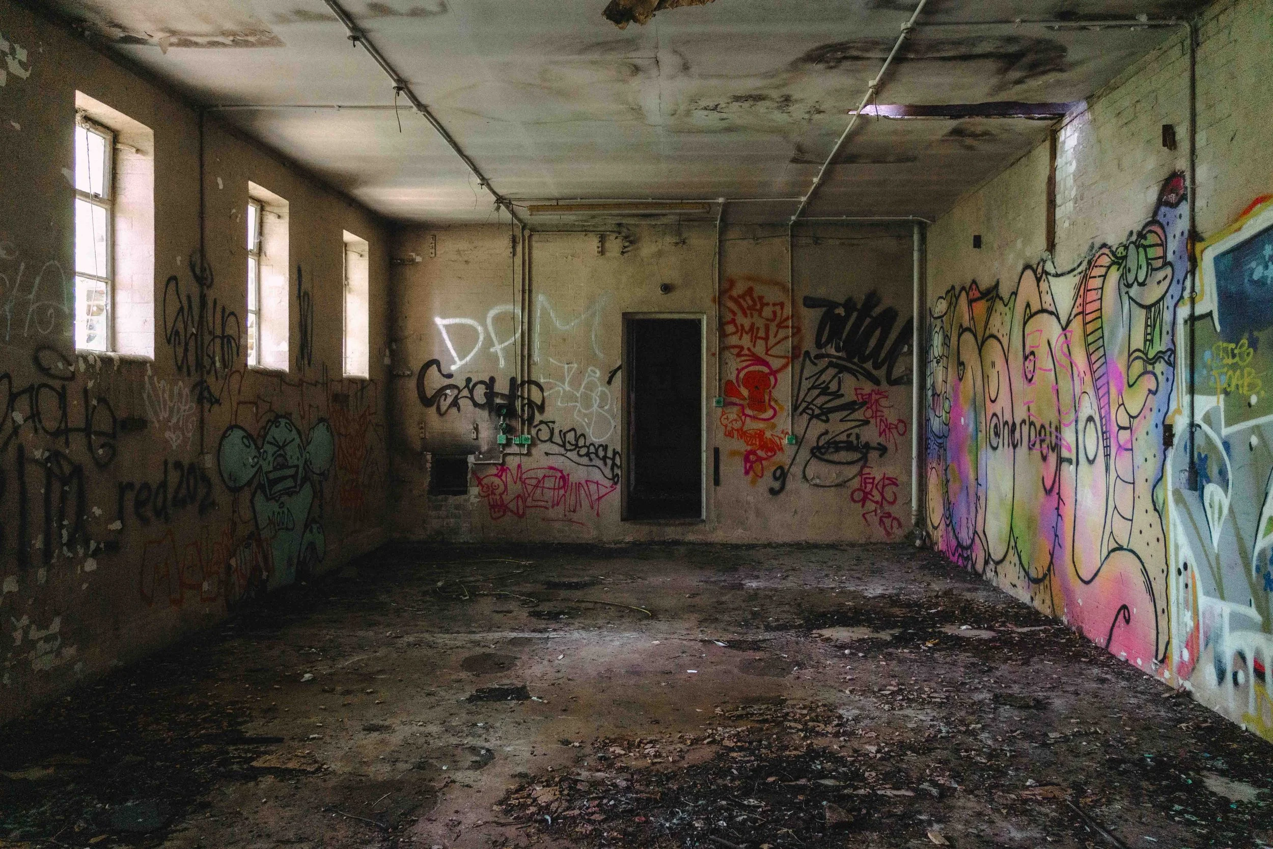 An abandoned, graffiti-covered room with a dirt floor, peeling walls, and three vents on the left wall. Natural light enters through multiple small windows on the left side. The ceiling has exposed pipes and signs of water damage.