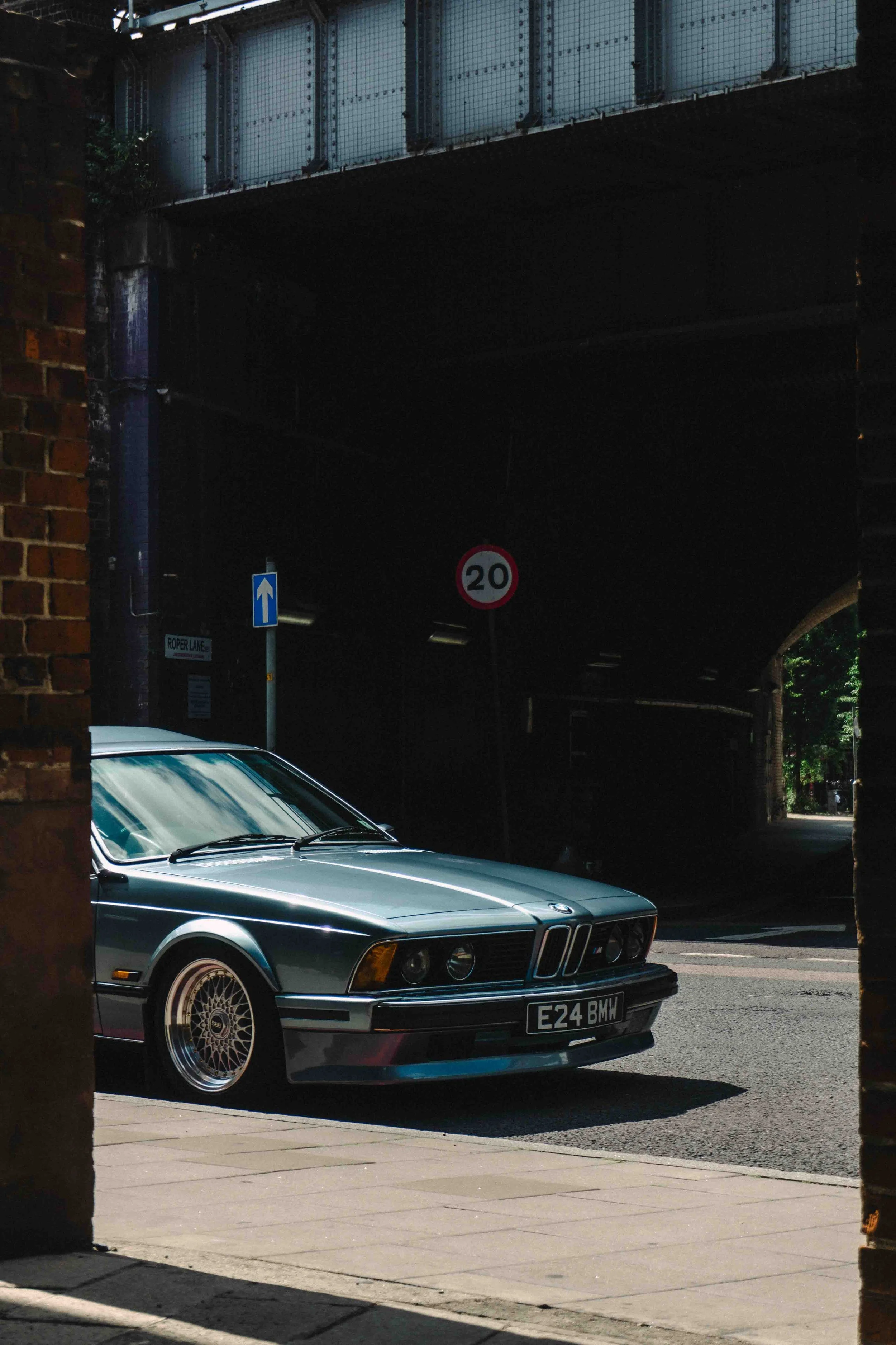 A vintage BMW E24 parked on a street, partially obscured by brick walls, with street signs and a bridge overhead in the background.