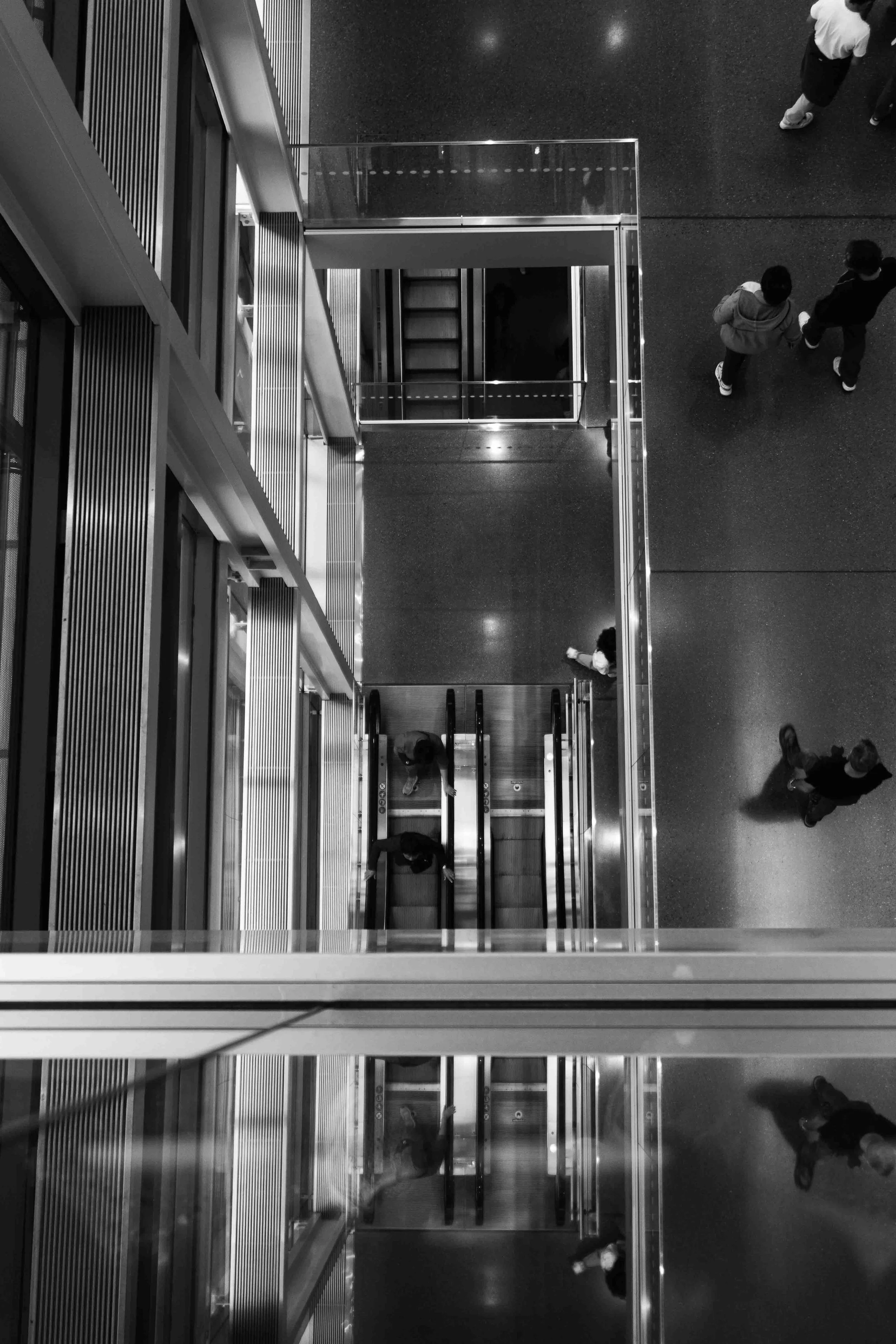 Black and white photo of an indoor multi-level building with an open elevator shaft, escalators, and people walking on the floors.