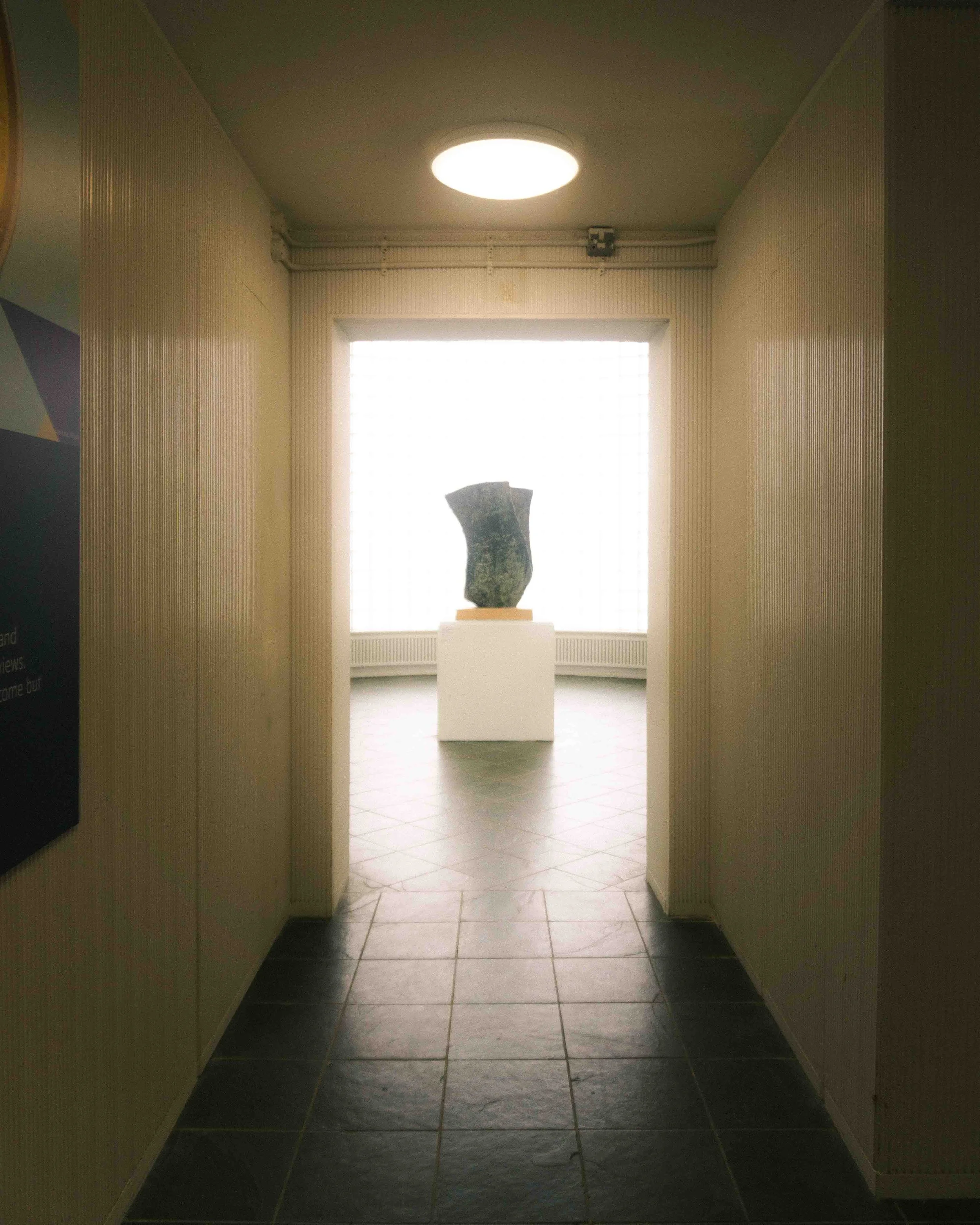 A hallway leading to an art exhibit room with a statue on a pedestal in front of a large window.