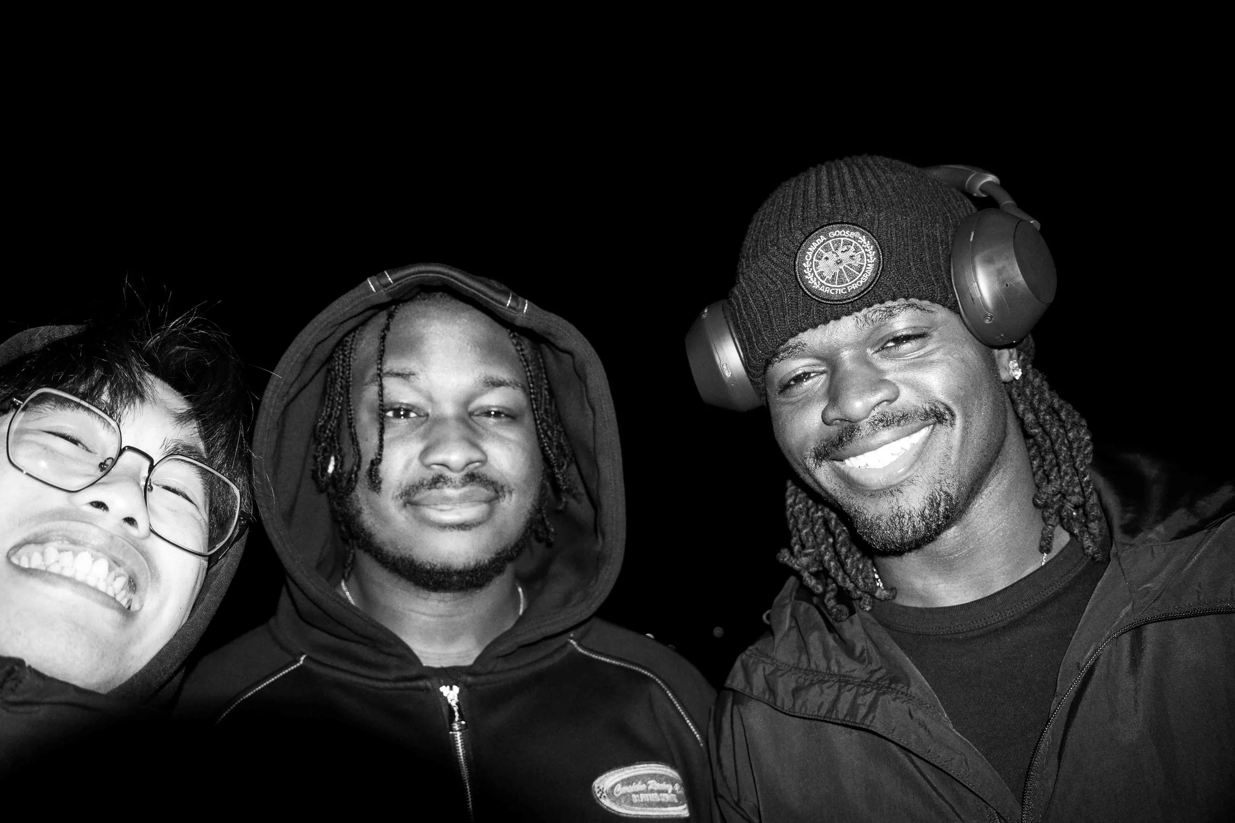 Three men smiling, posing closely together at night, one wearing glasses, one with braided hair and hoodie, one with dreadlocks, beanie, and headphones.