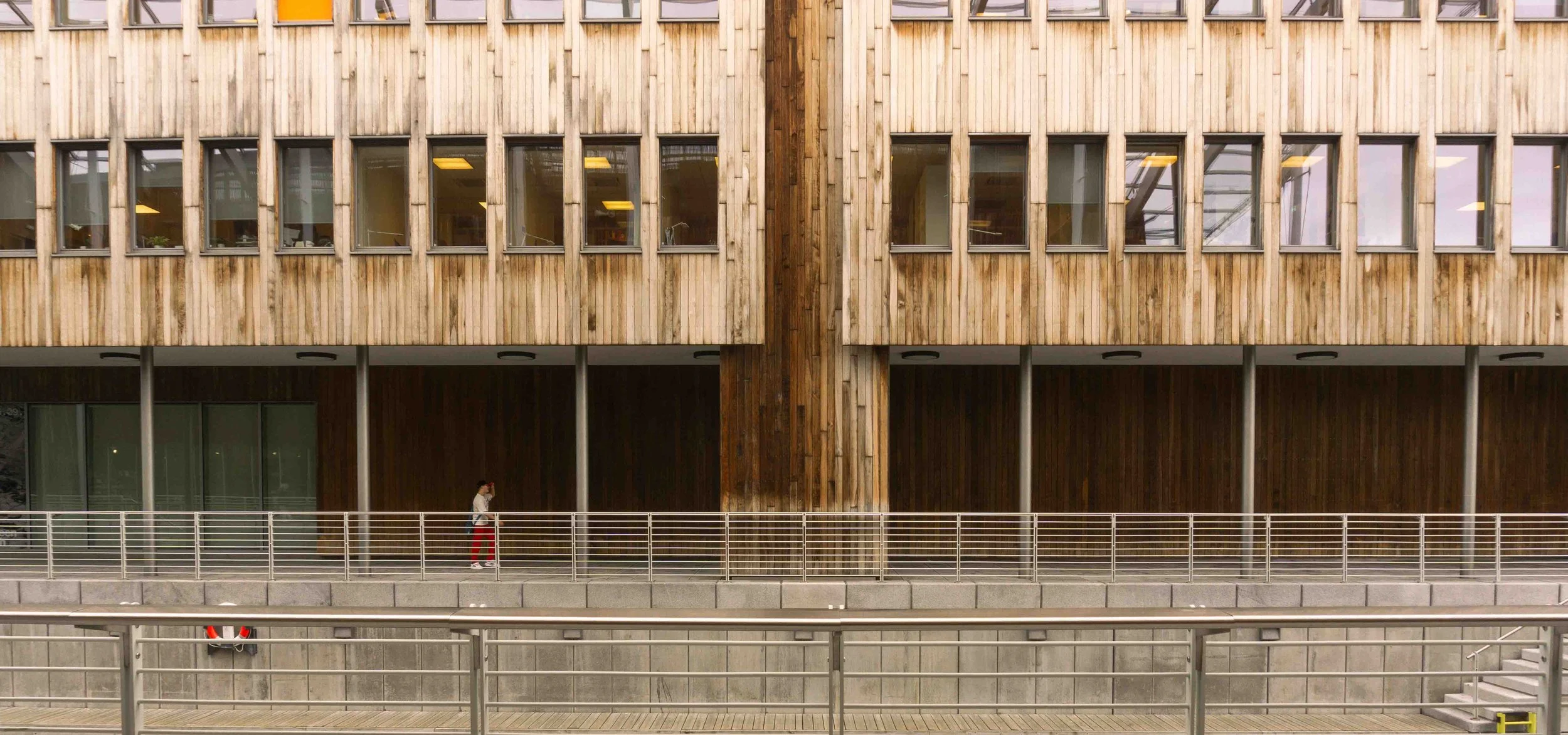 A person walking along a wooden-facade building with large windows, under a covered walkway, with metal railings and stairs nearby.