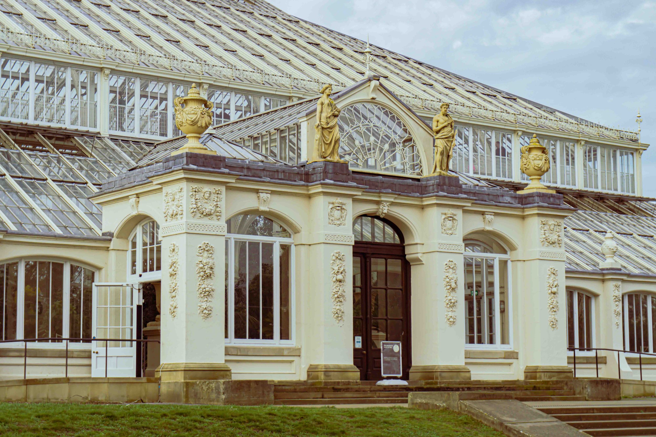 The image shows the exterior of a conservatory or greenhouse with ornate architectural details, including gold statues and decorative urns on top of the structure, and large glass windows and roof panels.