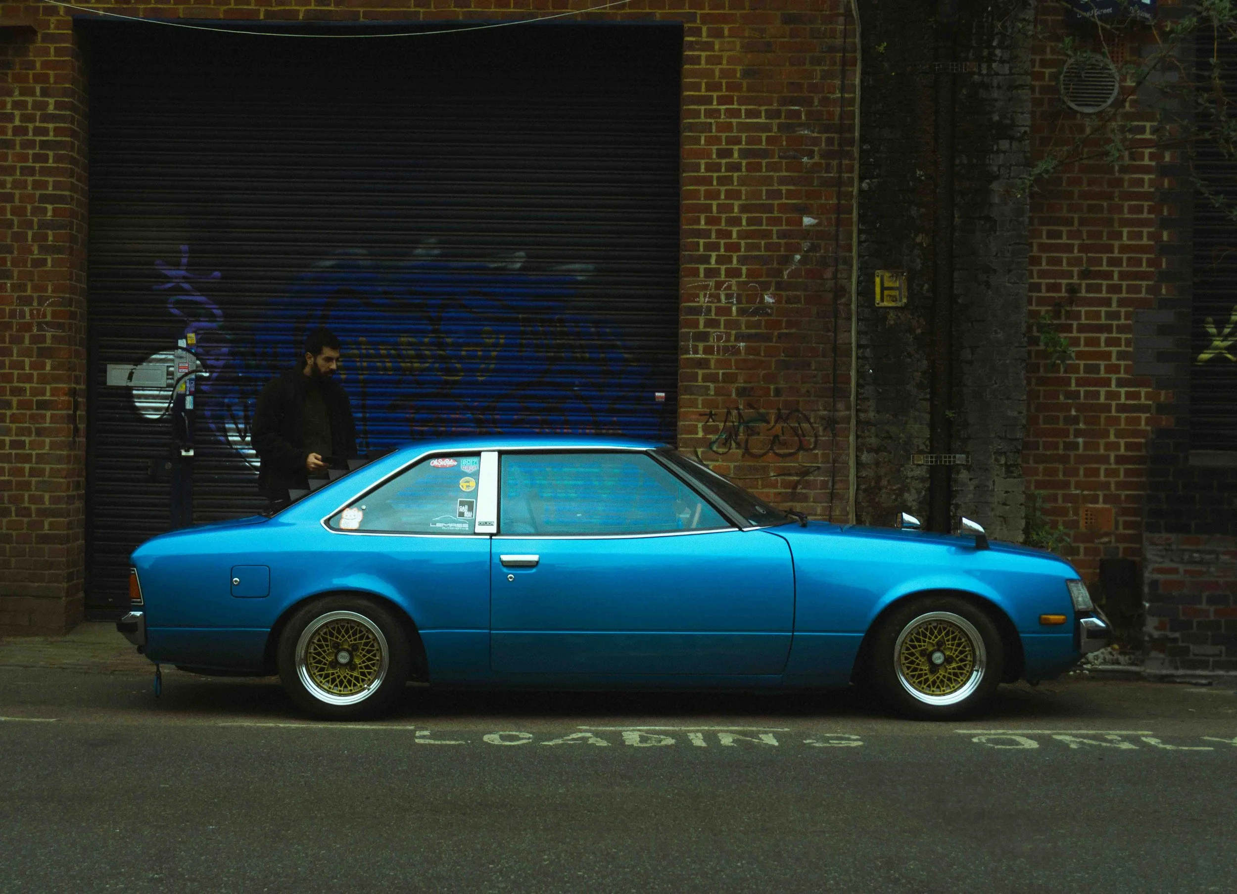 A man in a dark jacket standing behind a bright blue vintage sports car parked on a city street at night, with a brick building and graffiti in the background.