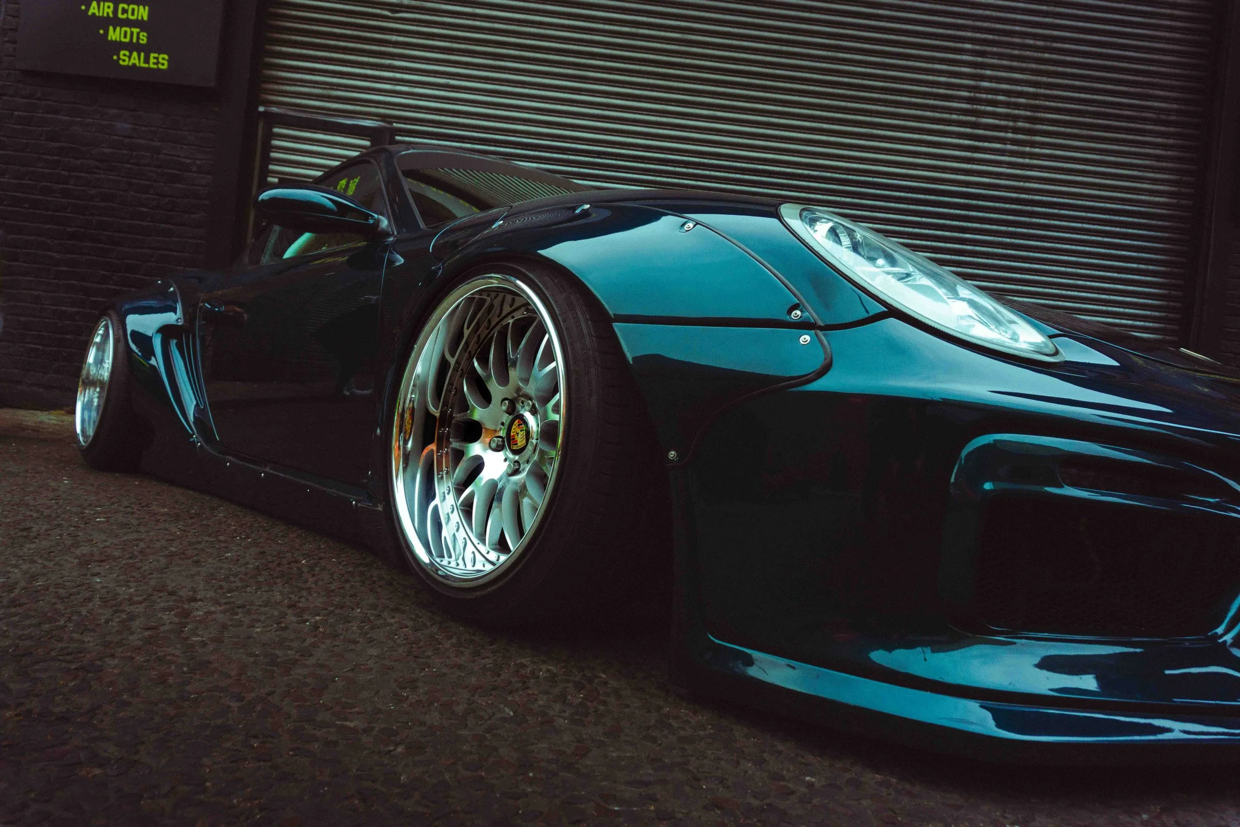 A sleek black sports car with large silver rims parked on a textured pavement, in front of a dark brick building with a metal rolling door.