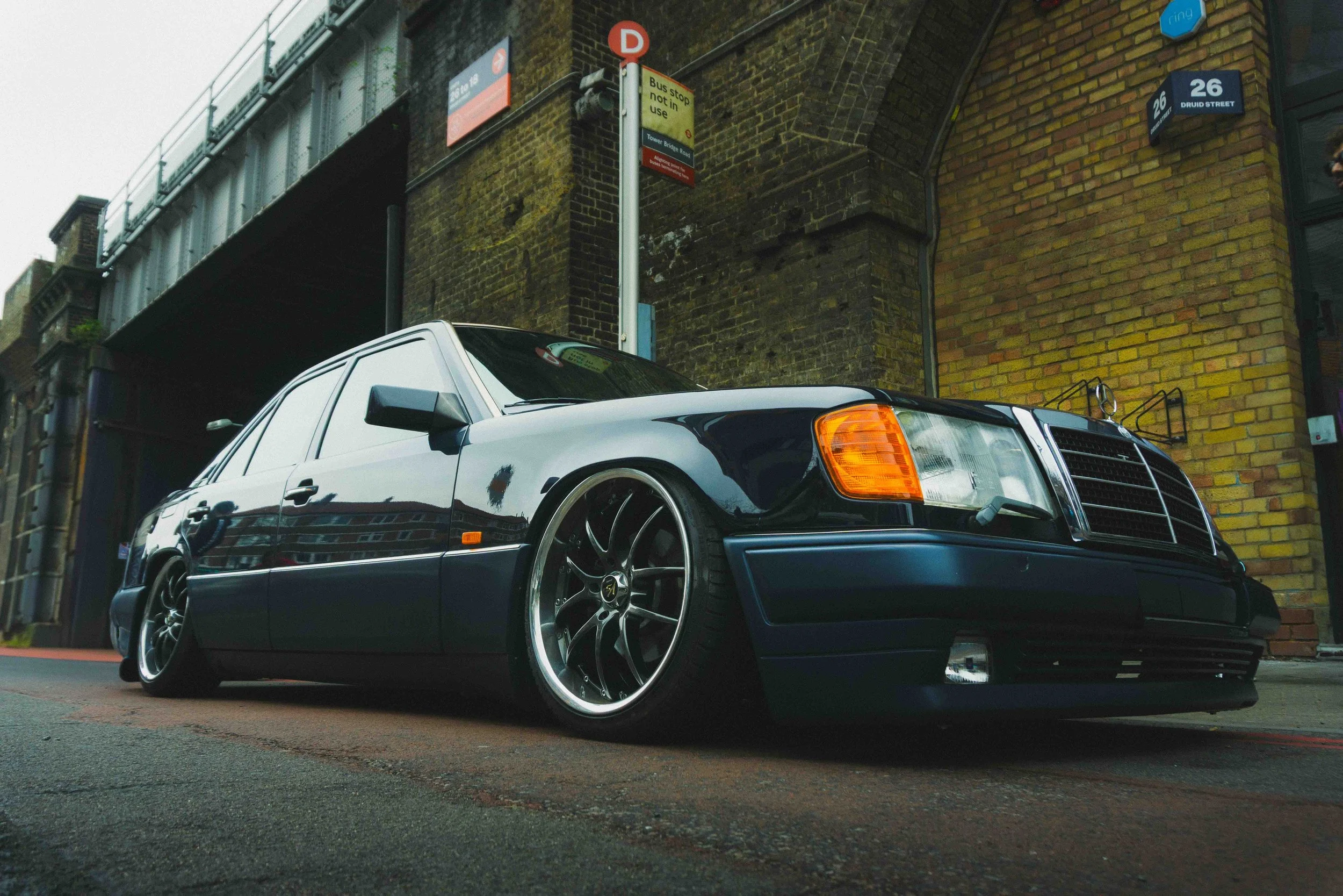 A lowered black Mercedes-Benz sedan with custom rims parked on a street, reflecting nearby buildings on its shiny surface. Behind it, there's a brick building with a yellow and brown facade and street signs, including a bus stop sign and a street add