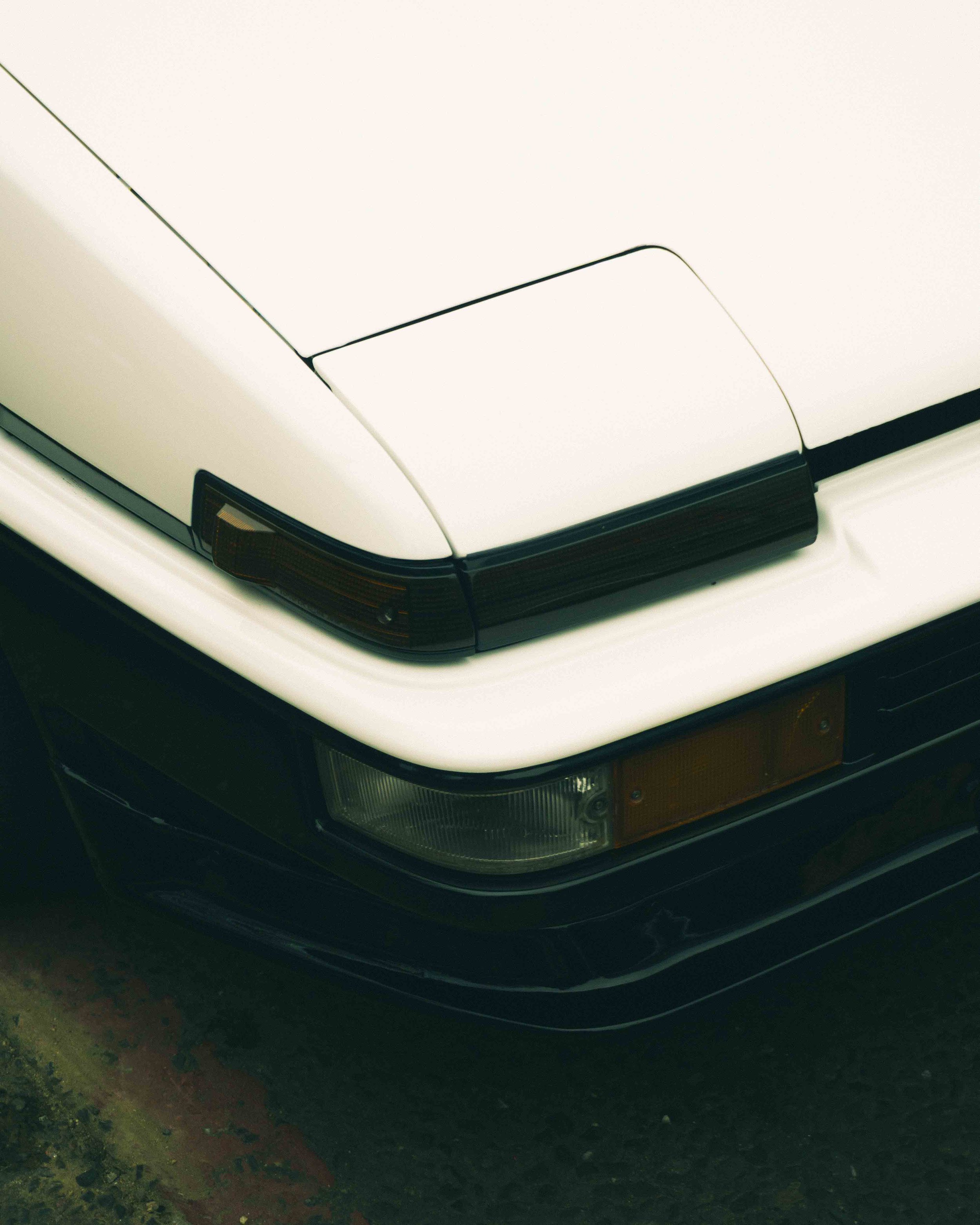 Close-up of the front corner of a white vintage car showing part of the hood, headlight, and turn signal.