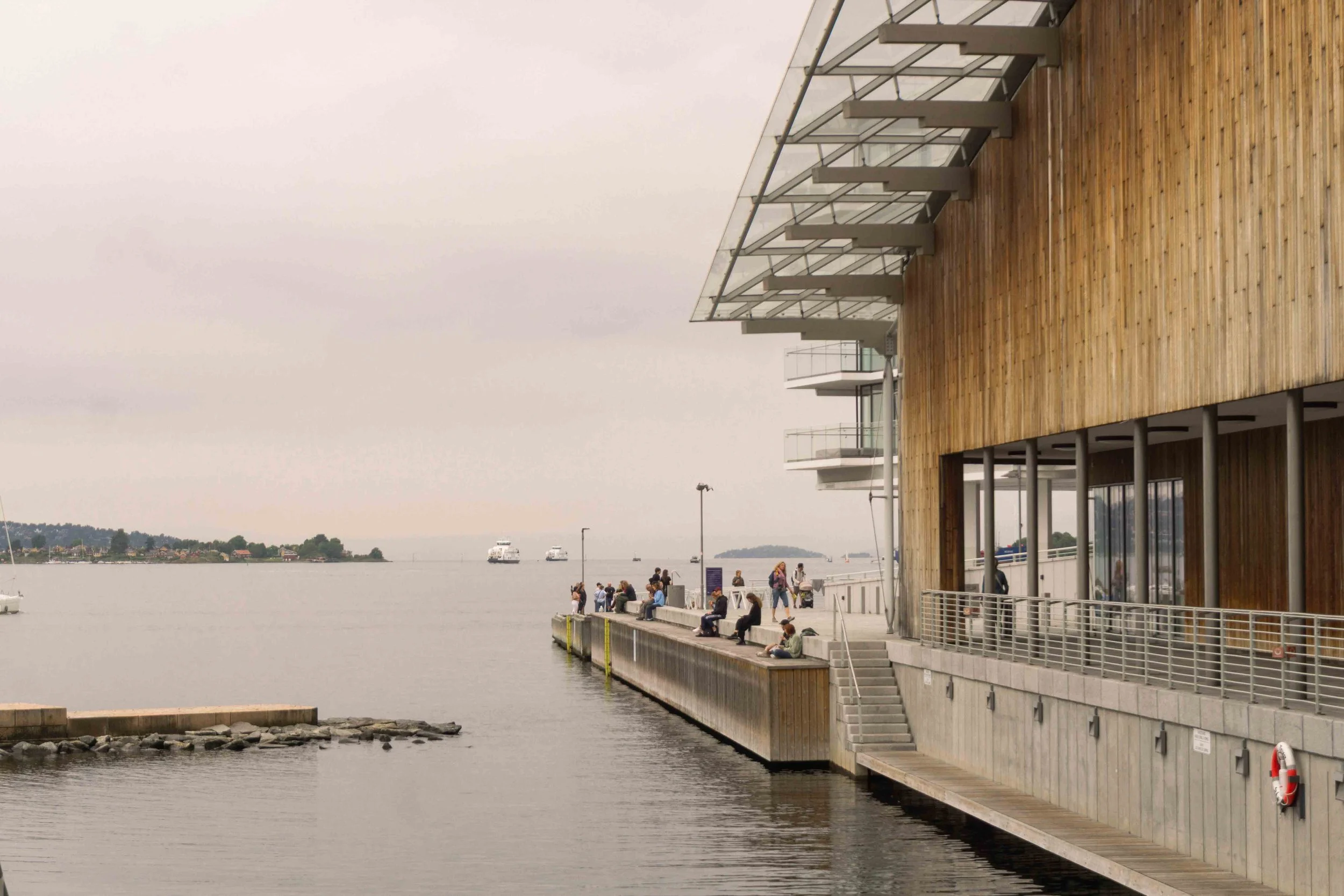 People sitting and standing on a pier next to a modern building with wooden exterior, overlooking the water with boats and a distant shoreline.