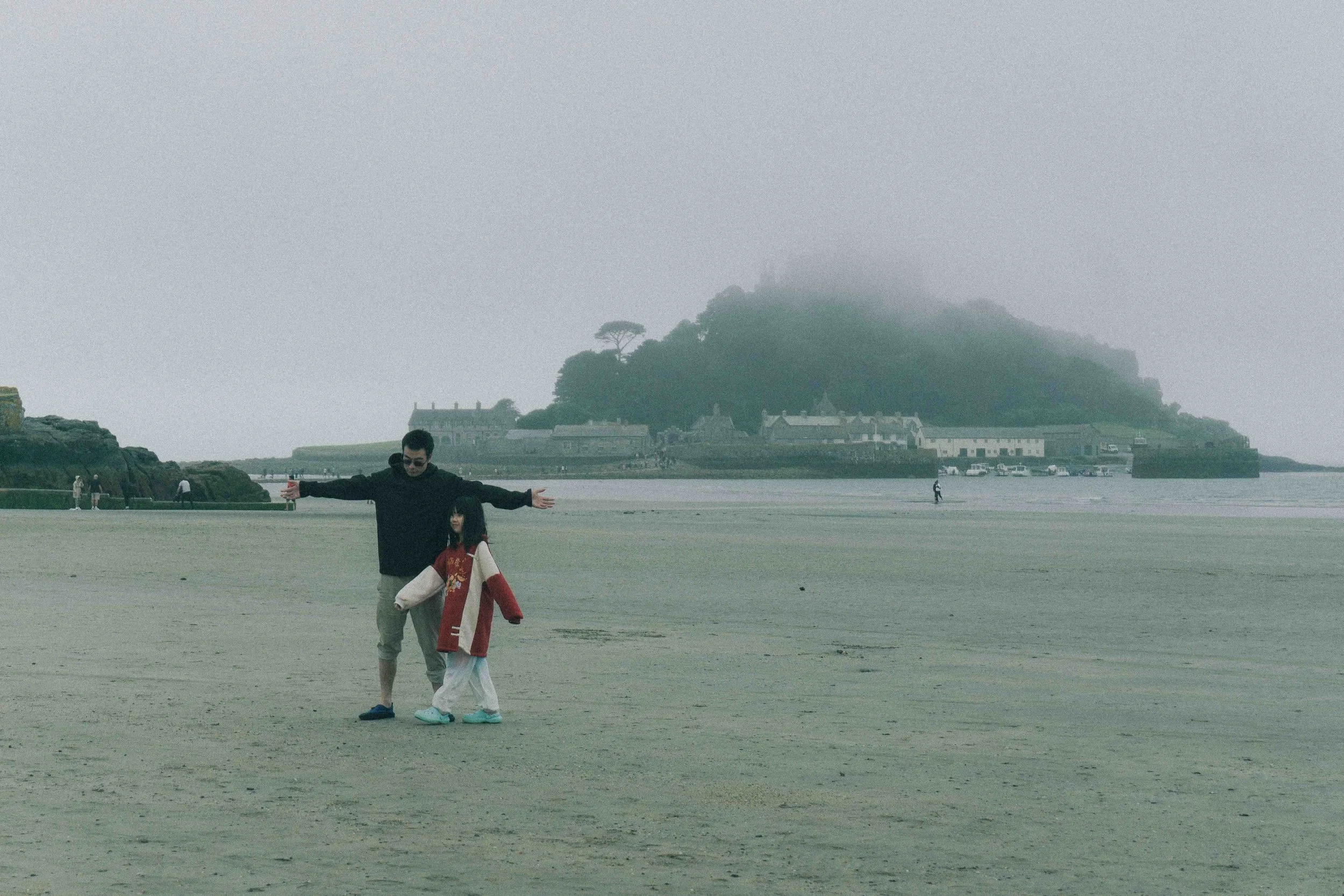 A man and a girl stand on a foggy beach with arms outstretched. In the background, there is a hill with fog and some buildings and trees on it, along with a few other people walking along the beach and some cars parked near the buildings.