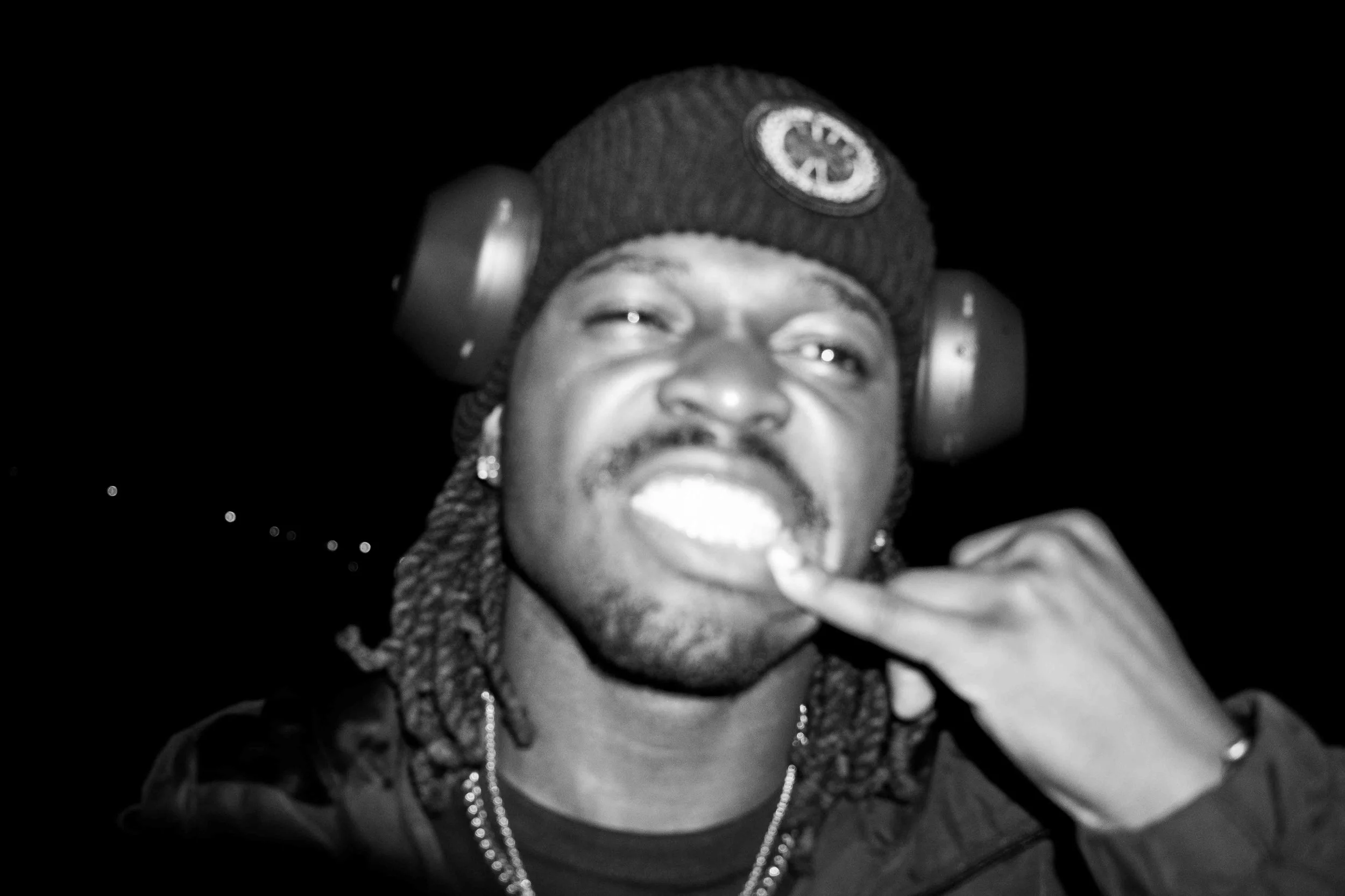 A young man with dreadlocks wearing a beanie with a logo, headphones, and a chain, showing his teeth and making a gesture with his hand against a dark background.