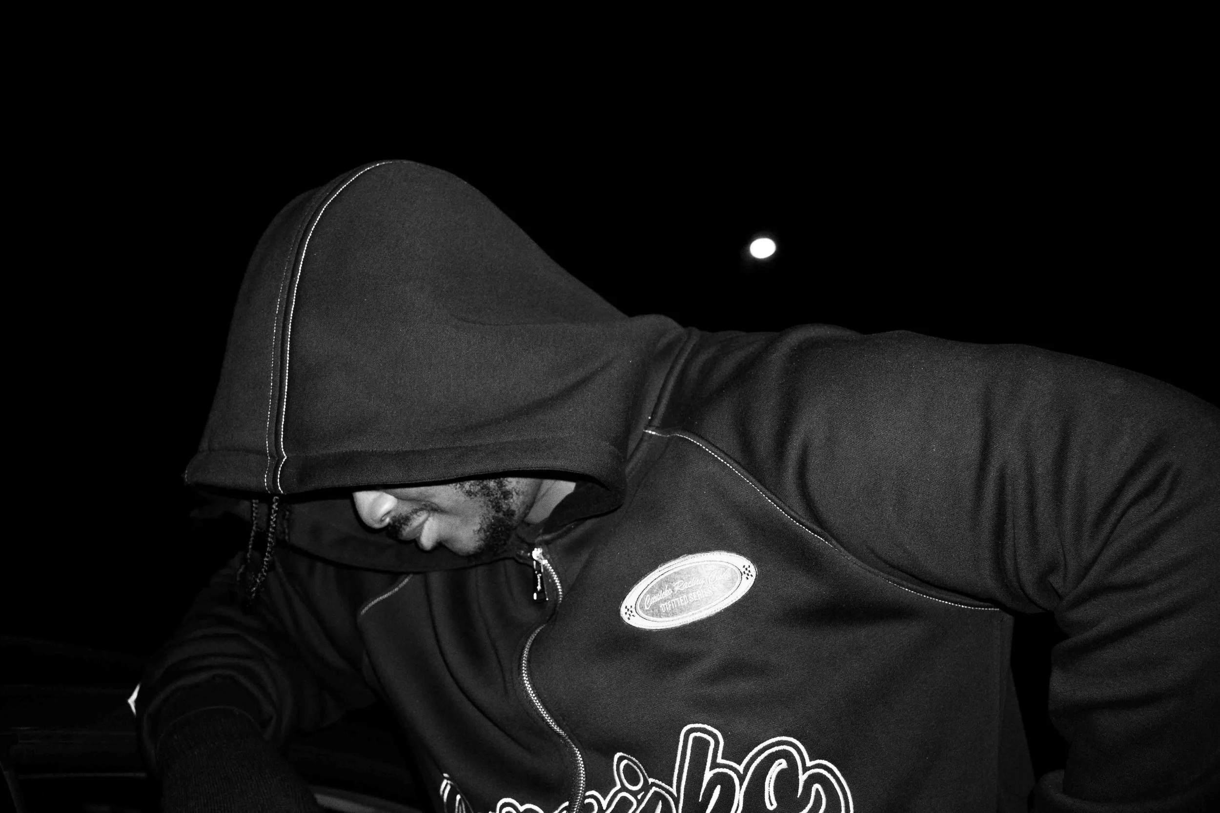 A man wearing a hoodie with the hood over his head, looking down, outside at night. The moon is visible in the dark sky behind him.
