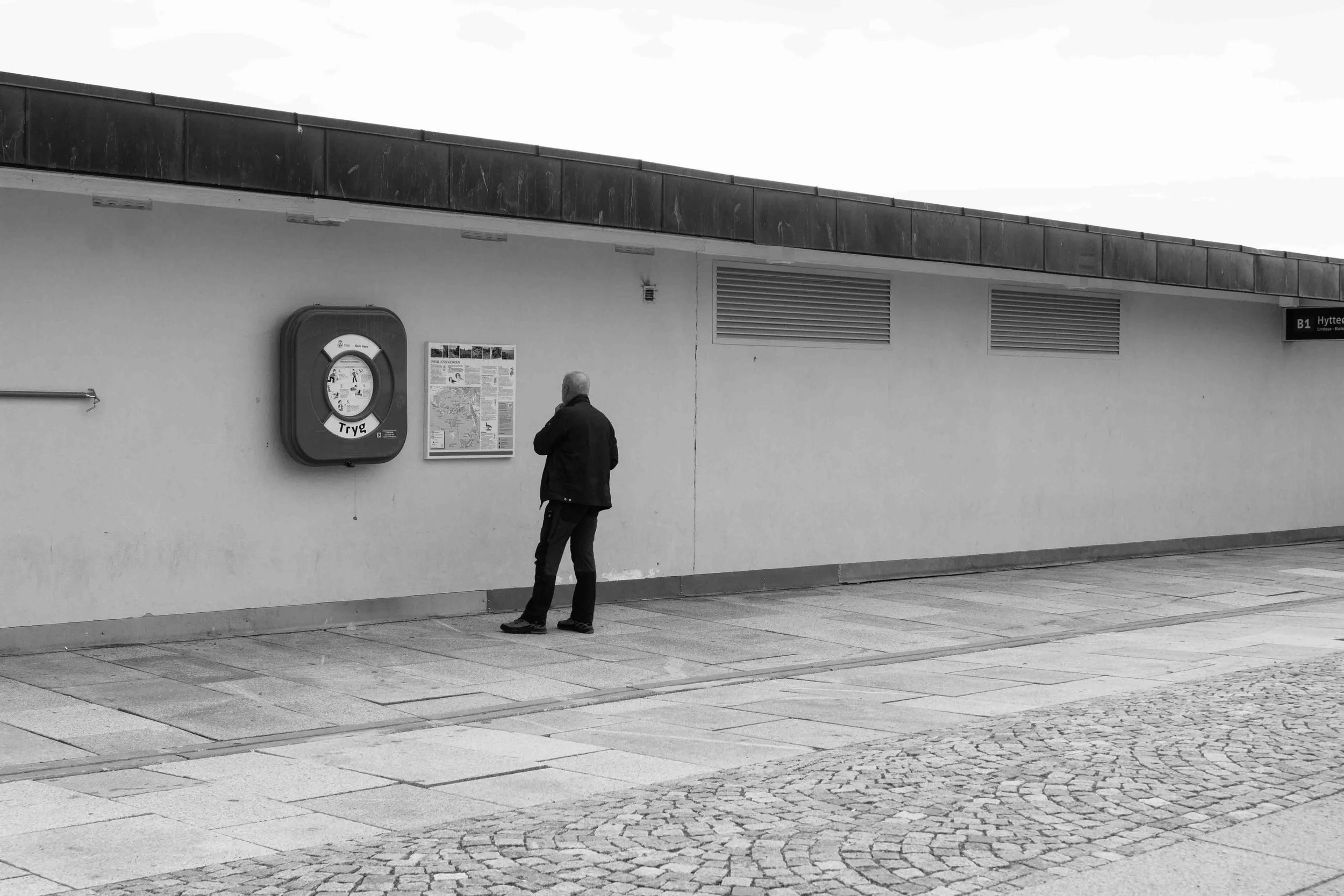 A man in a dark jacket and pants standing on a sidewalk in front of a plain wall, looking at a notice board.
