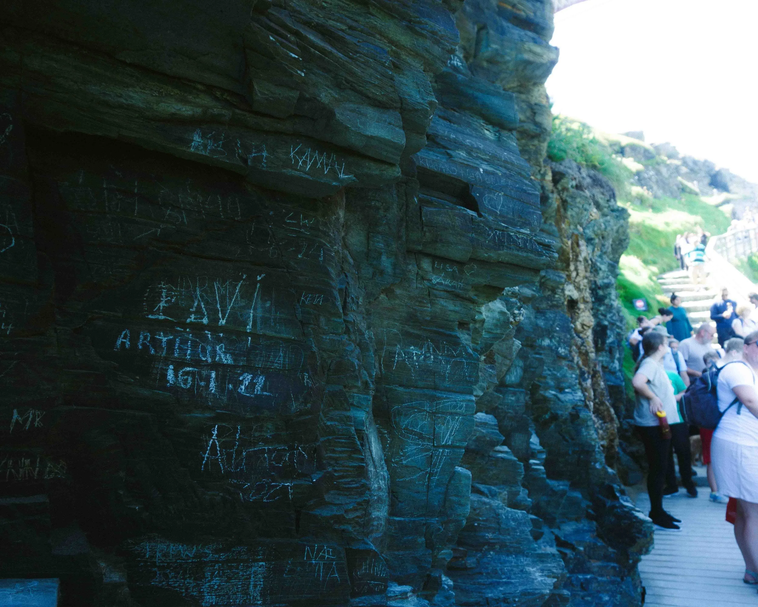 People walking on a trail with rock formations on the left and a green landscape in the background.