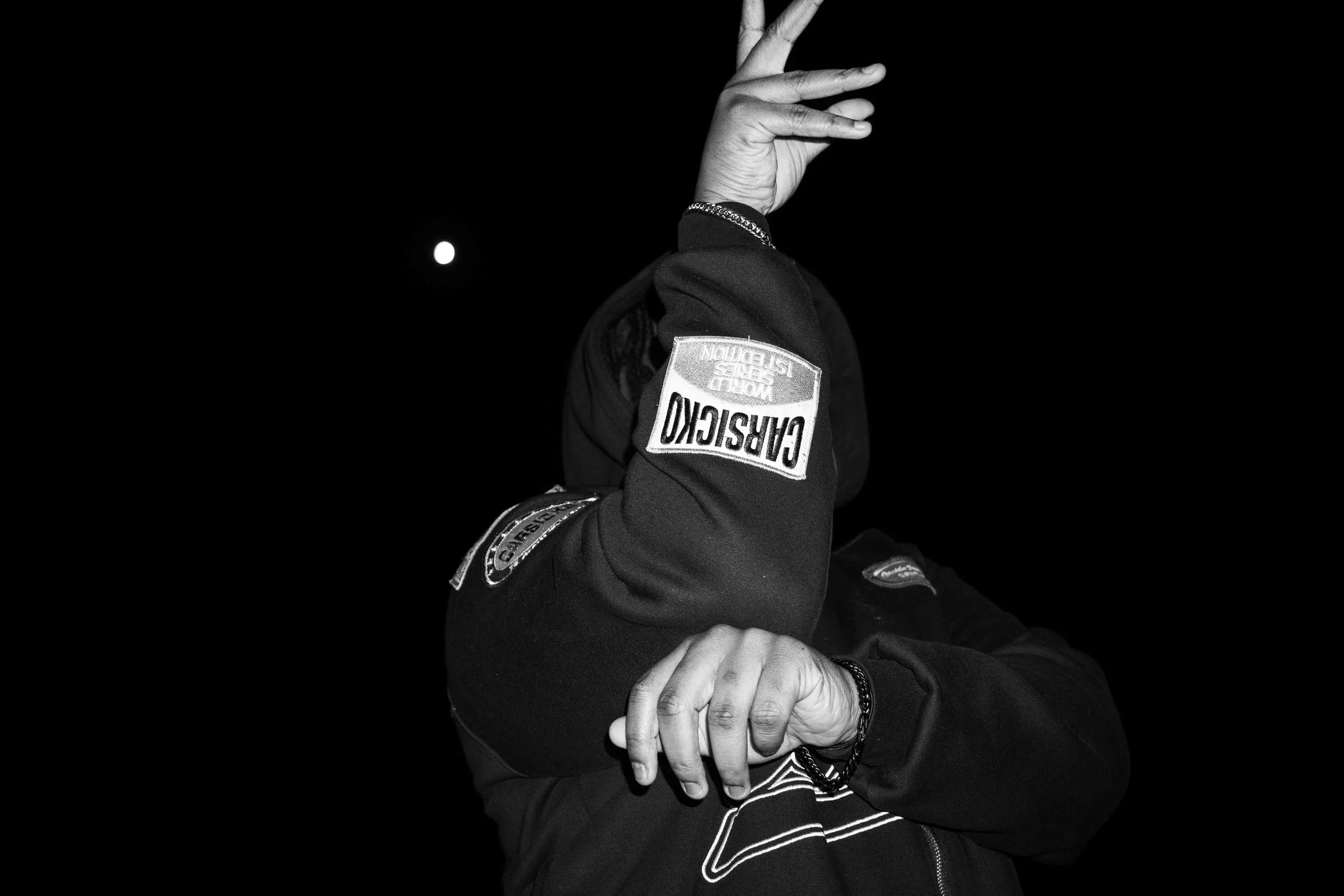 A person in a dark jacket with patches, covering their face with one arm and hand, against a dark background with a small bright object, possibly the moon, in the sky.