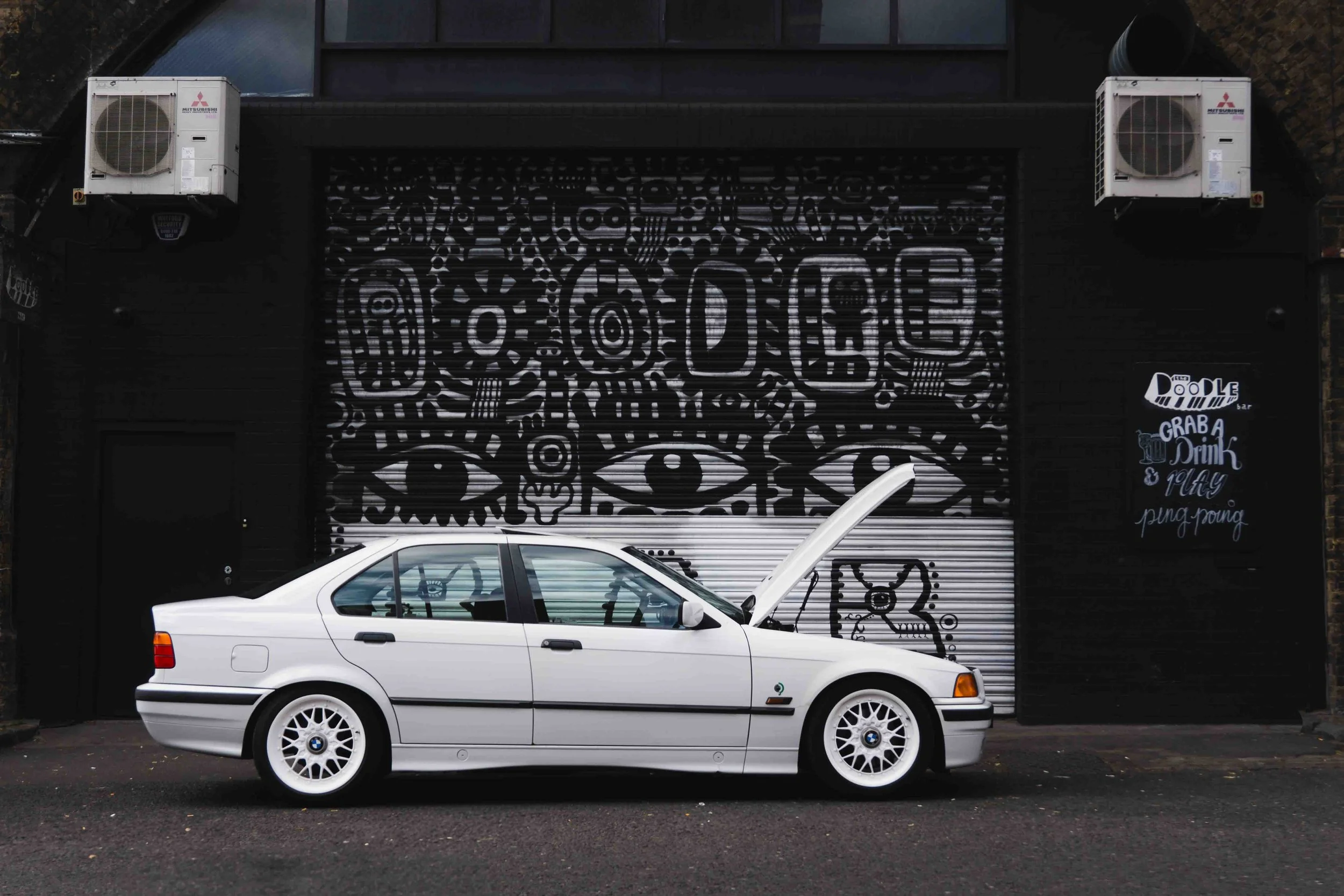 A white BMW car with the hood open parked in front of a black building with street art. The street art features large eyes, abstract patterns, and the word 'DIEG' in capital letters. There are two air conditioning units mounted on the building's uppe
