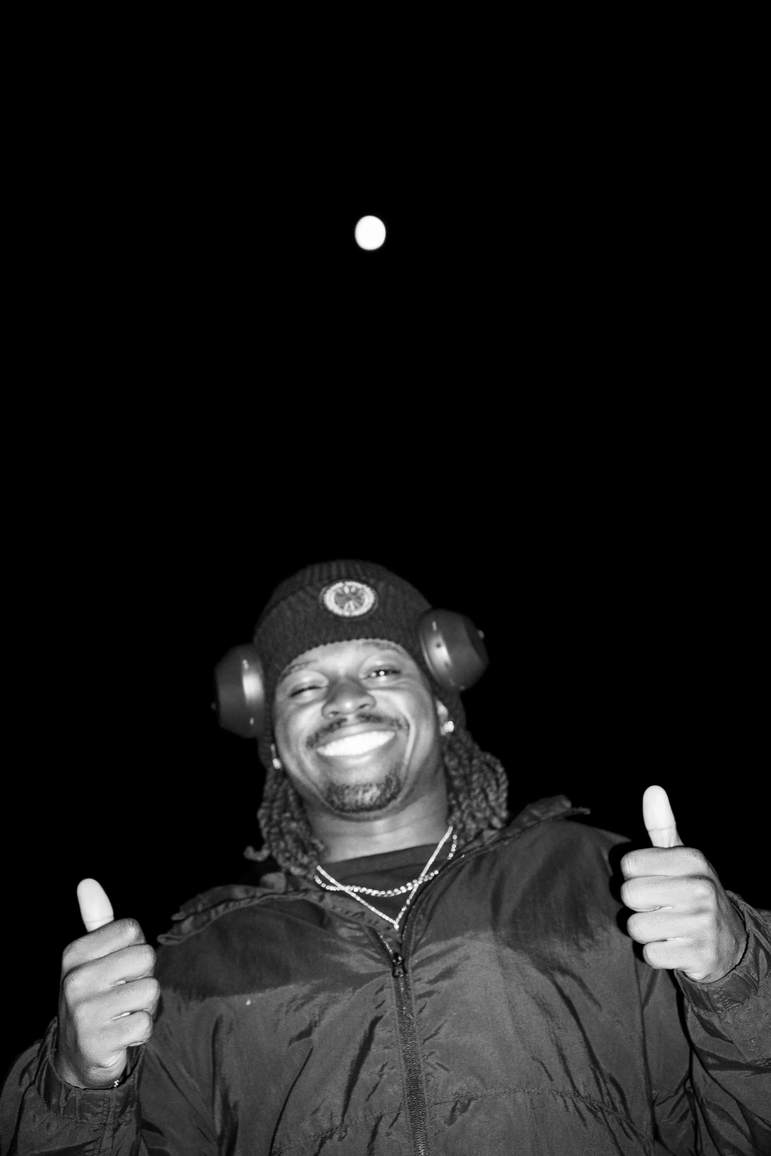 A smiling man with dreadlocks wearing a beanie, headphones, and a jacket, giving two thumbs up against a black night sky with a bright moon.