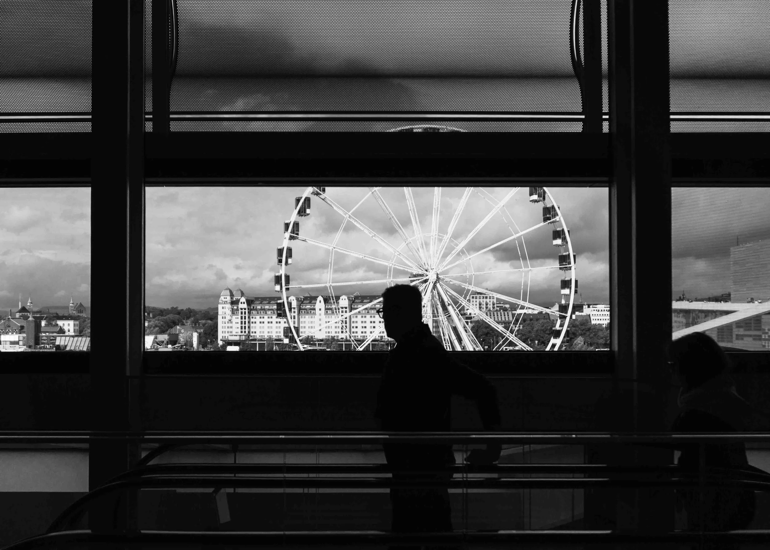 Black-and-white photo of a view through airport terminal window showing a large Ferris wheel outside, with two silhouetted people walking in the foreground.