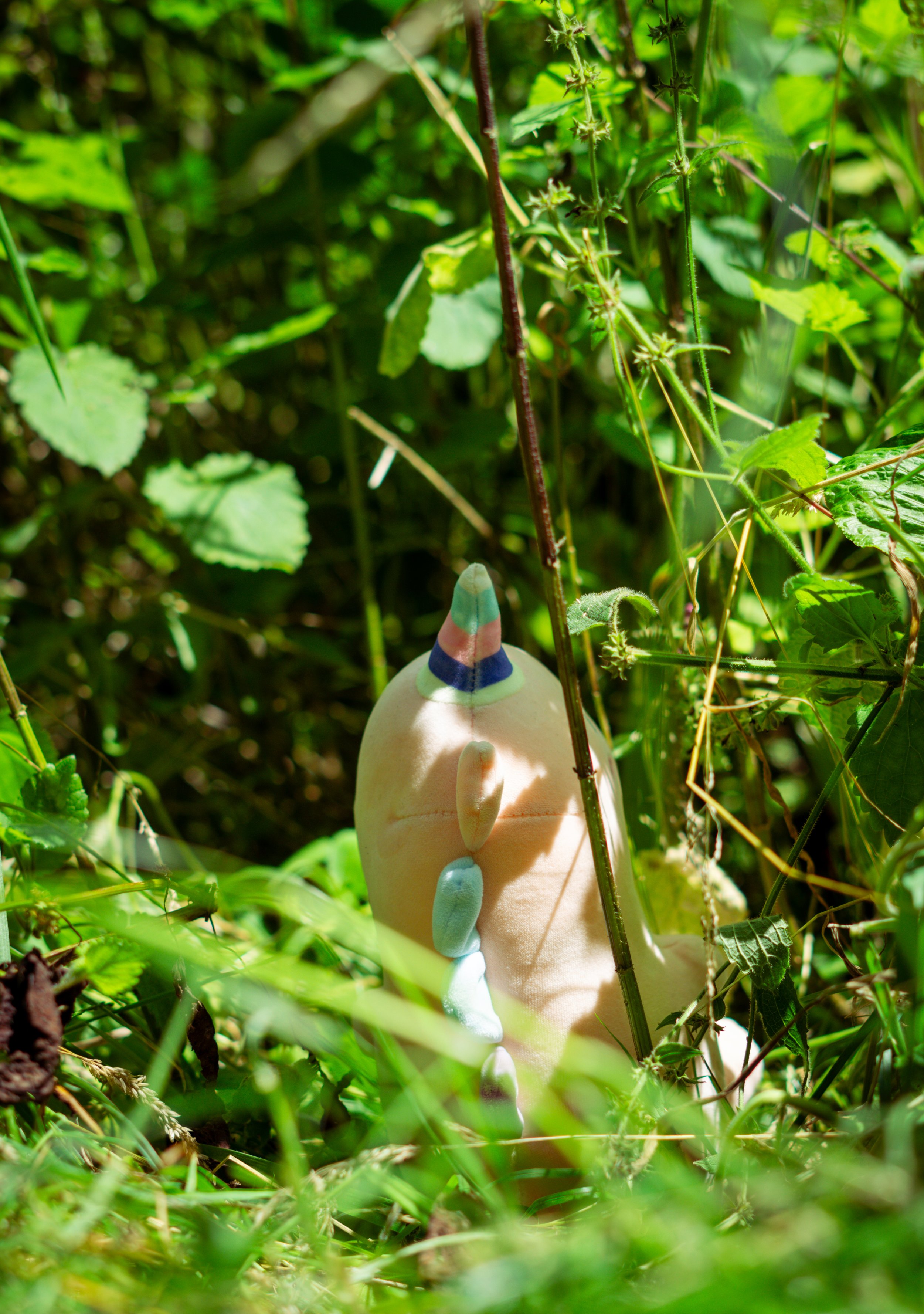 A colorful plastic toy unicorn partially hidden among green grass and leafy plants in a natural outdoor setting.
