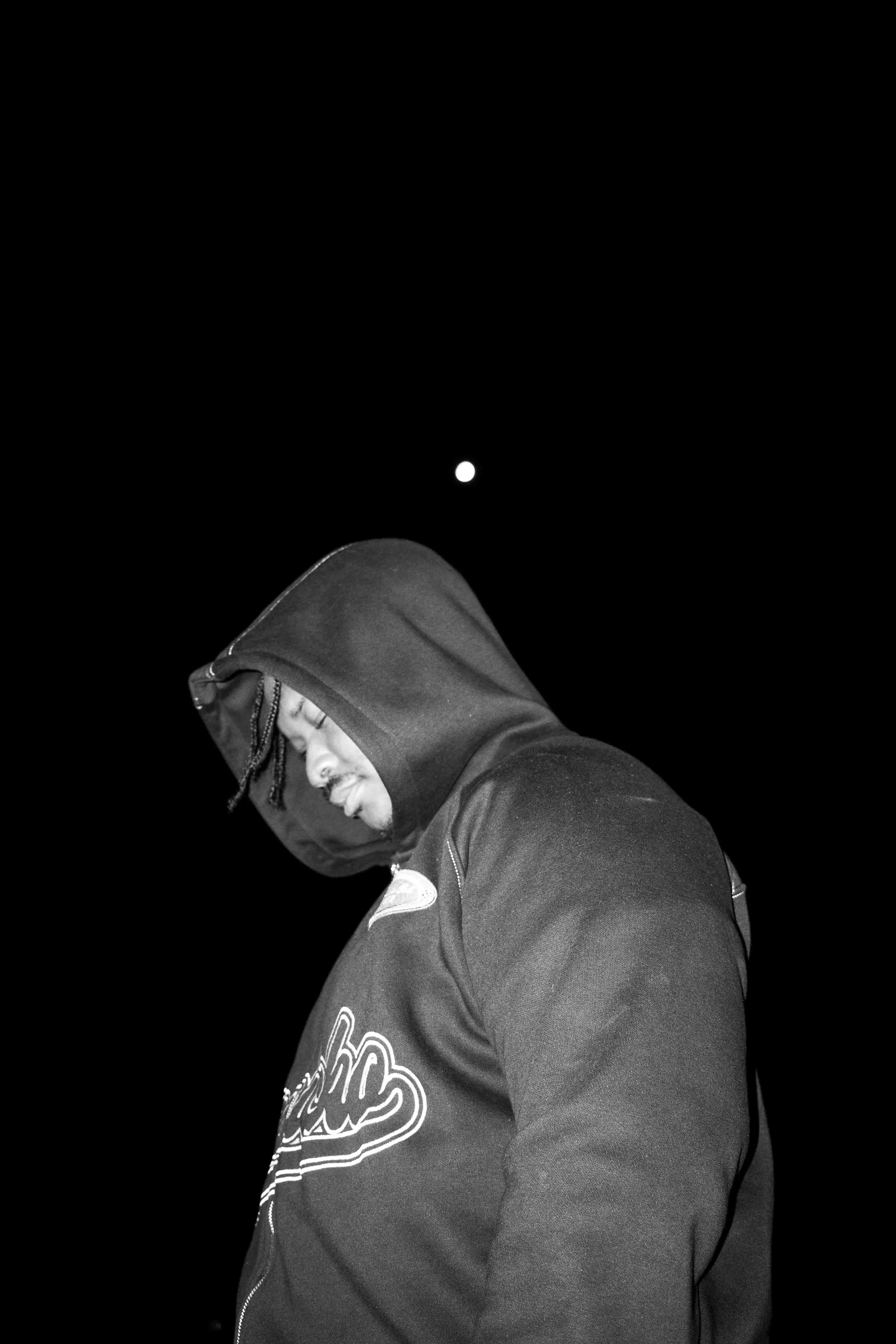Black and white photo of a young man wearing a hoodie and jacket, looking down, with the moon visible in the dark night sky.