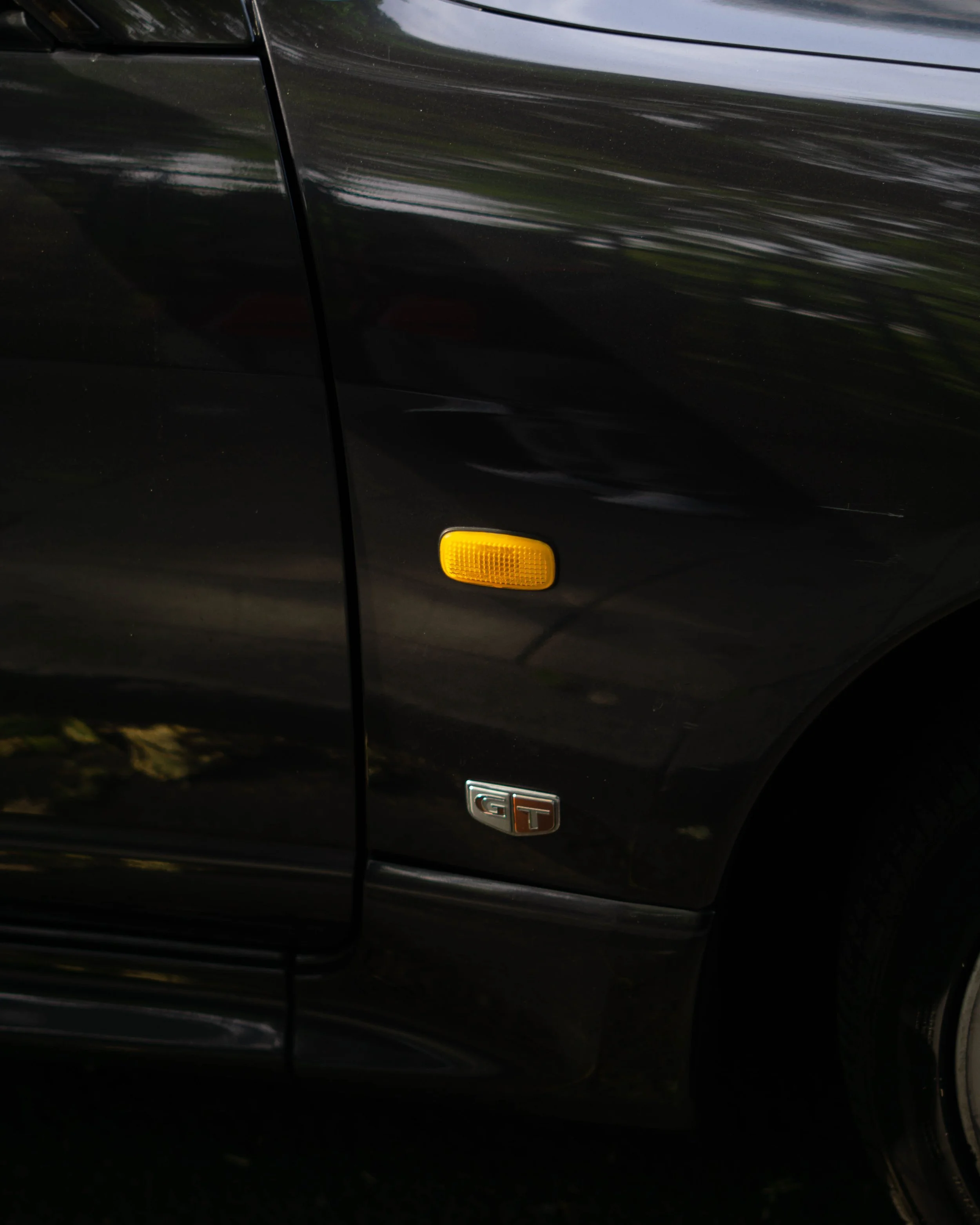Close-up of a black sports car's front side, featuring a yellow side marker light and a GT badge.
