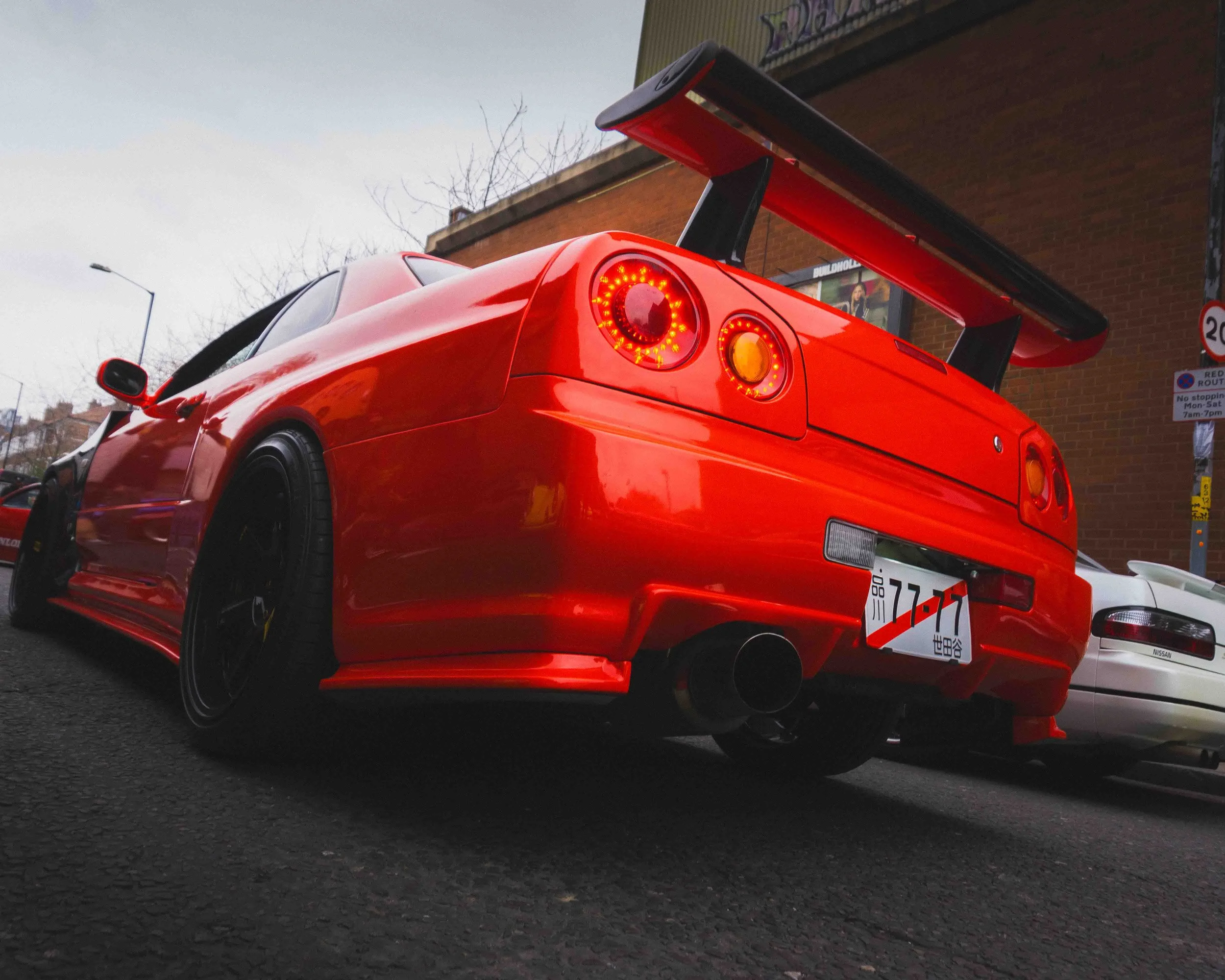 A red sports car with a large rear wing, black wheels, and an aftermarket exhaust, parked on an urban street.