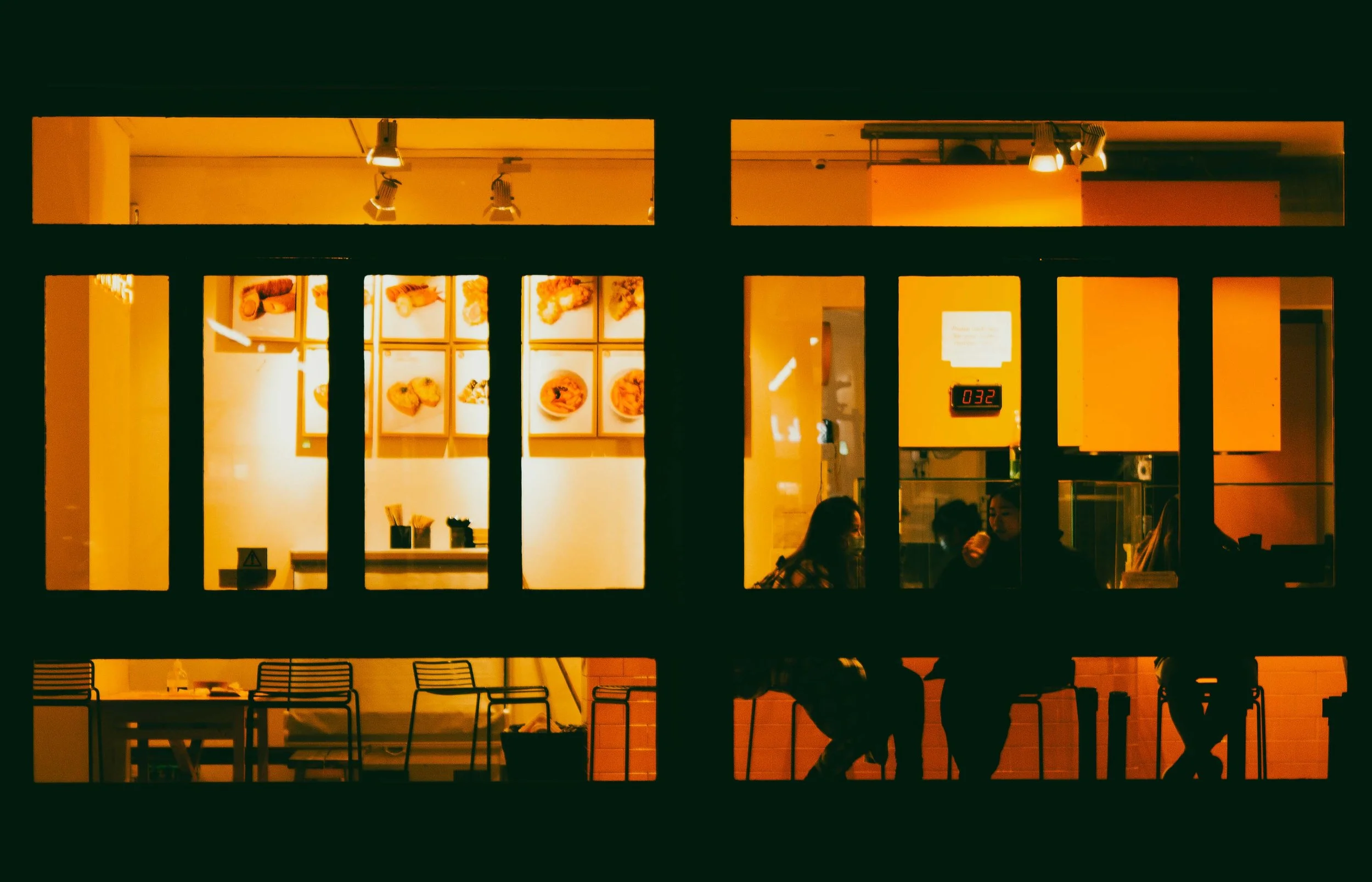 Night view of a café or restaurant seen through a window with black framing, showing interior with people, menu images on the wall, and warm yellow lighting.