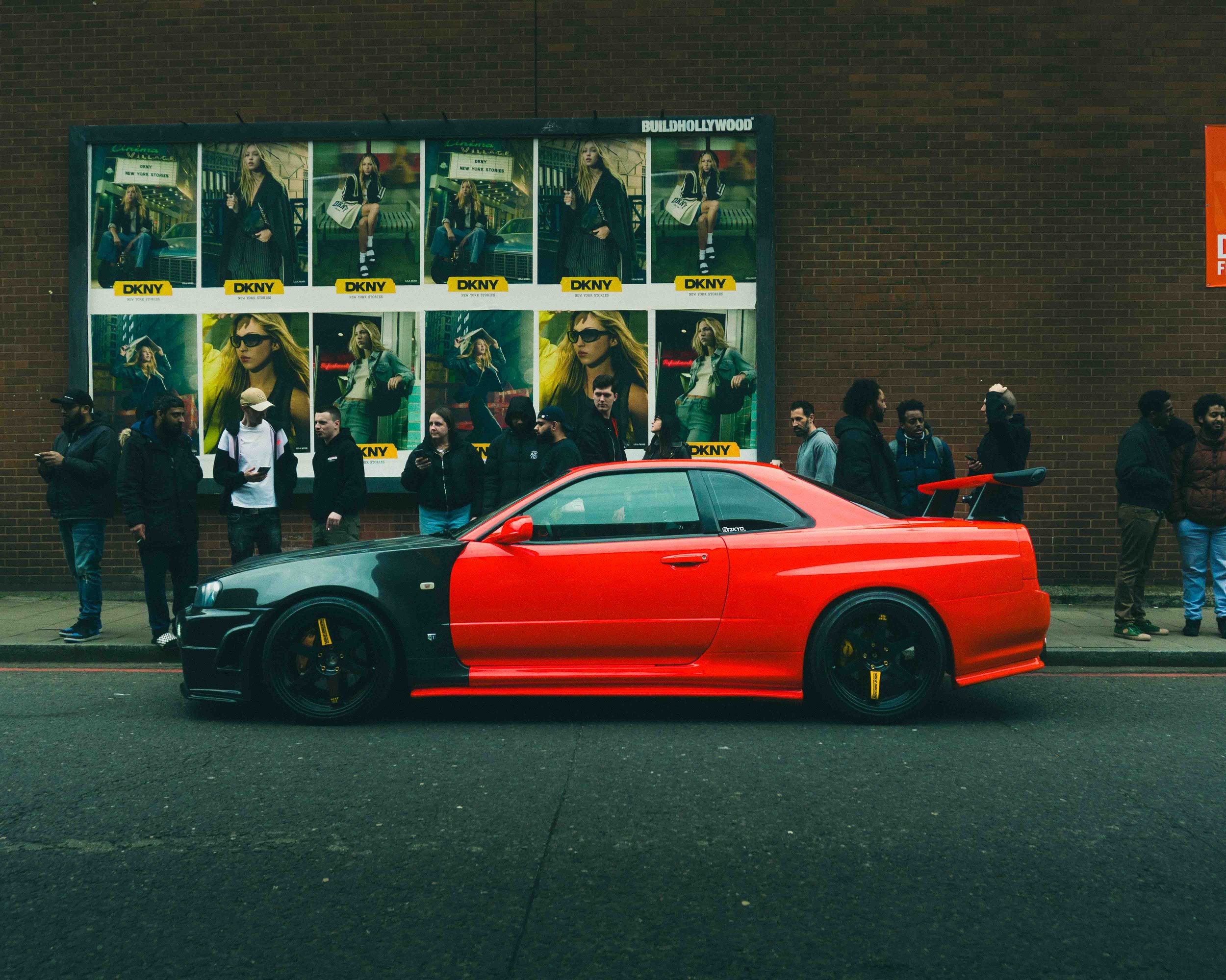 Red and black sports car parked on a city street with pedestrians and a brick wall with fashion advertisements in the background.