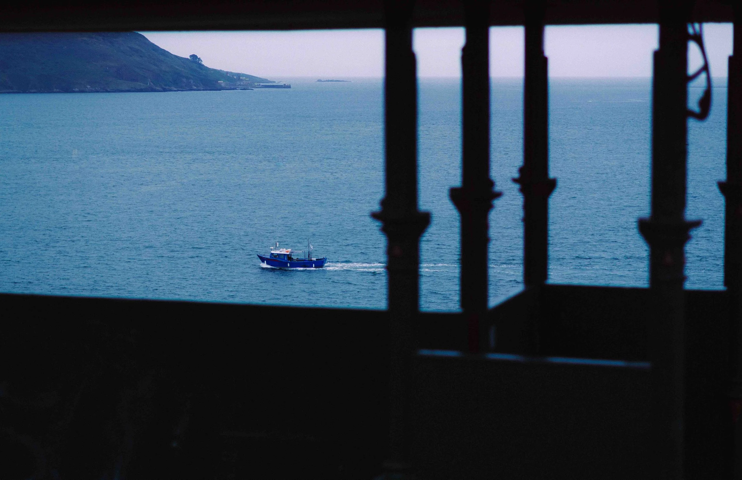 View of a blue boat sailing in the ocean, seen through a window with black metal bars, with land and hillside in the background.