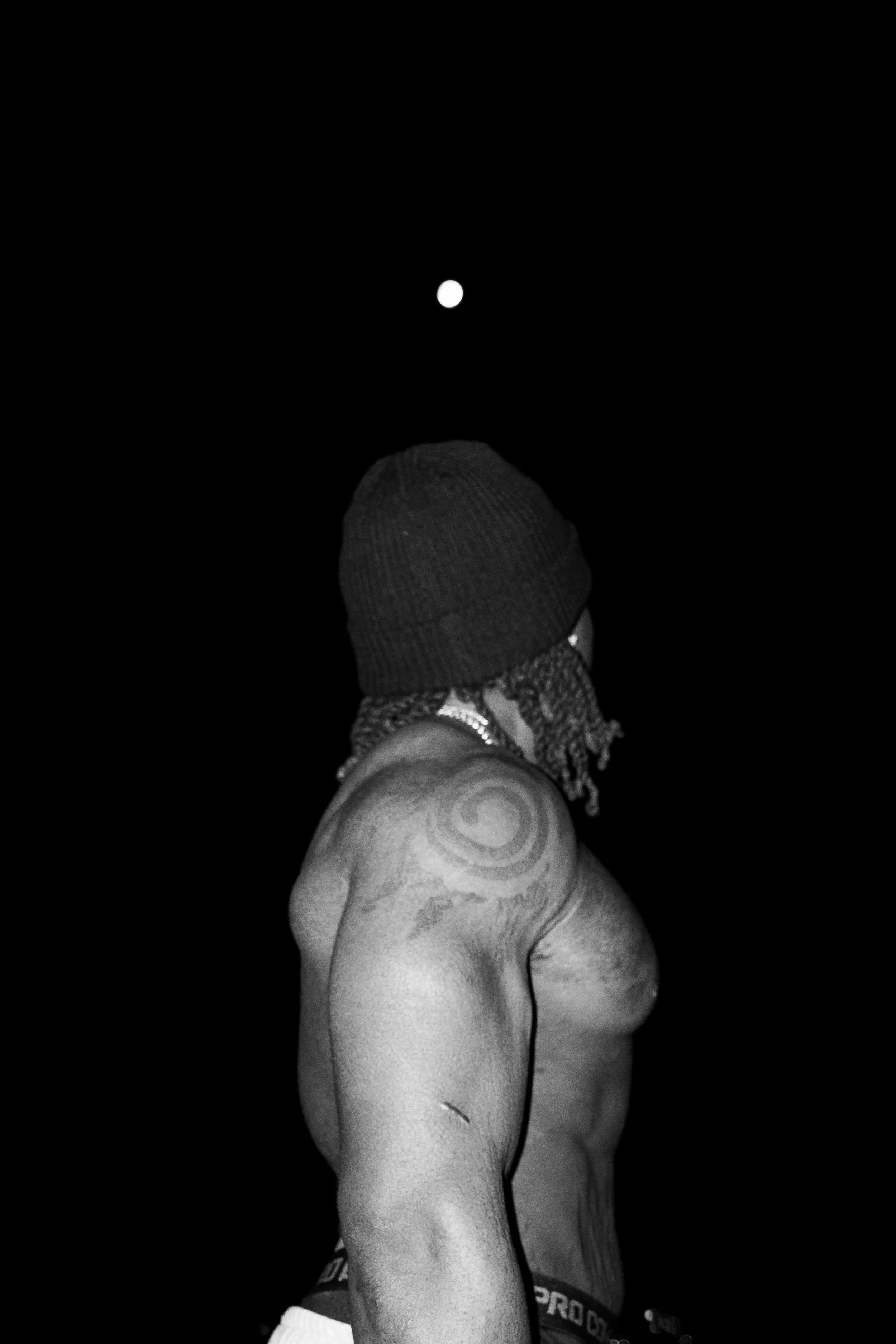 A shirtless man with dreadlocks, wearing a knit beanie and a chain necklace, standing against a black background with a full moon, showing a tattoo on his shoulder.