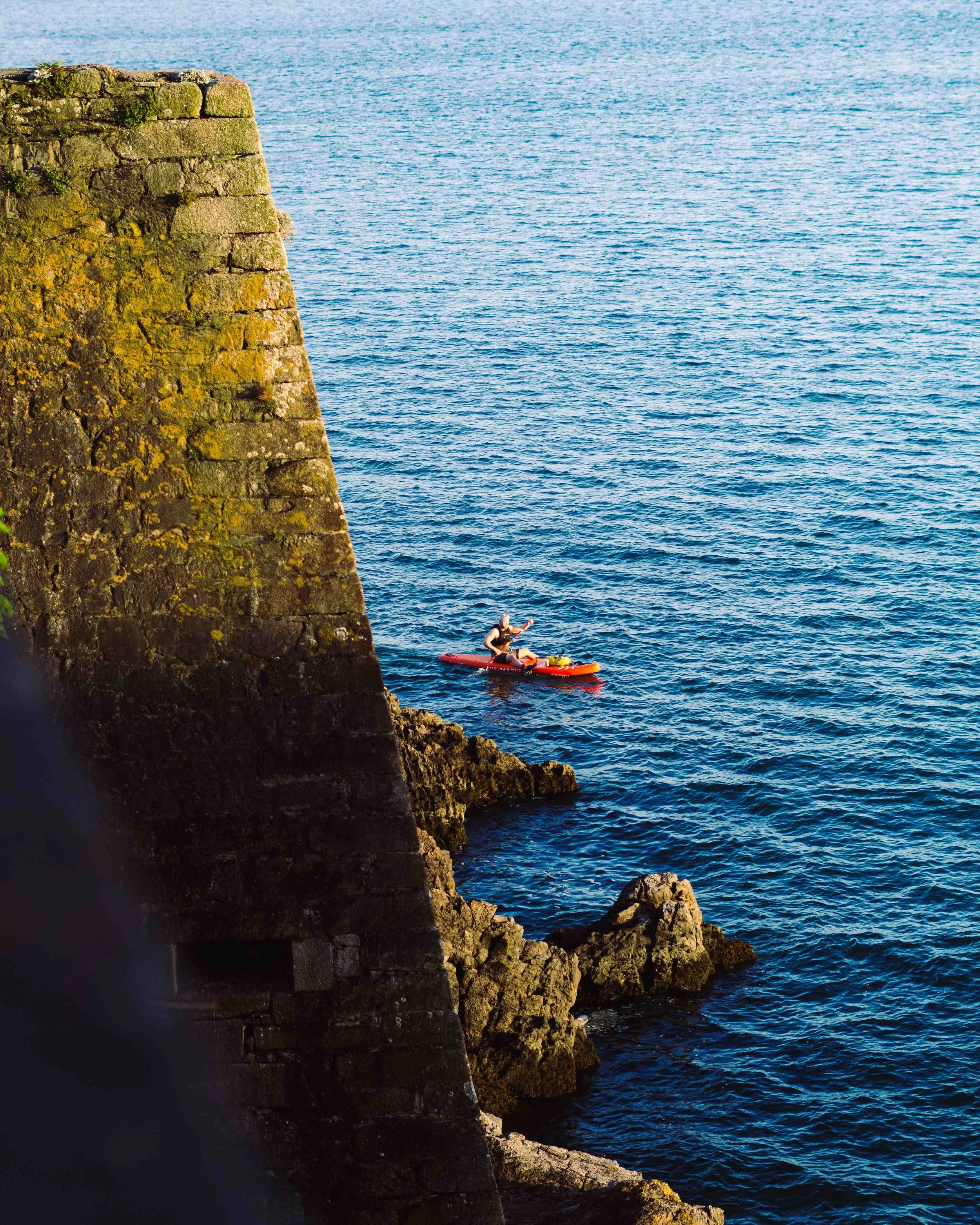 A person kayaking near a rocky shoreline and a large moss-covered stone wall.