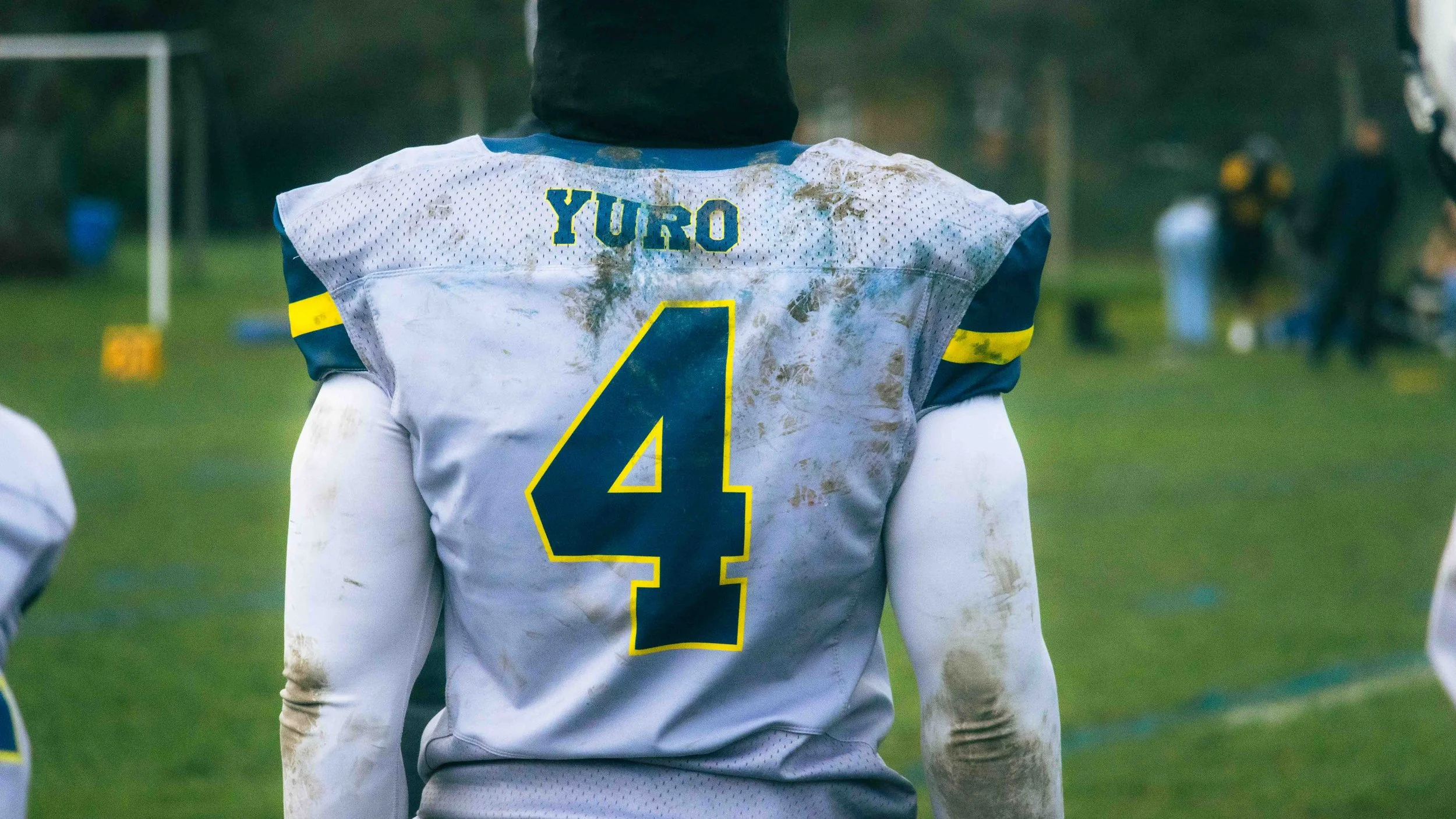 Back of a football player in a white uniform with the name 'Yuro' and the number 4, with dirt stains, on a grassy field.