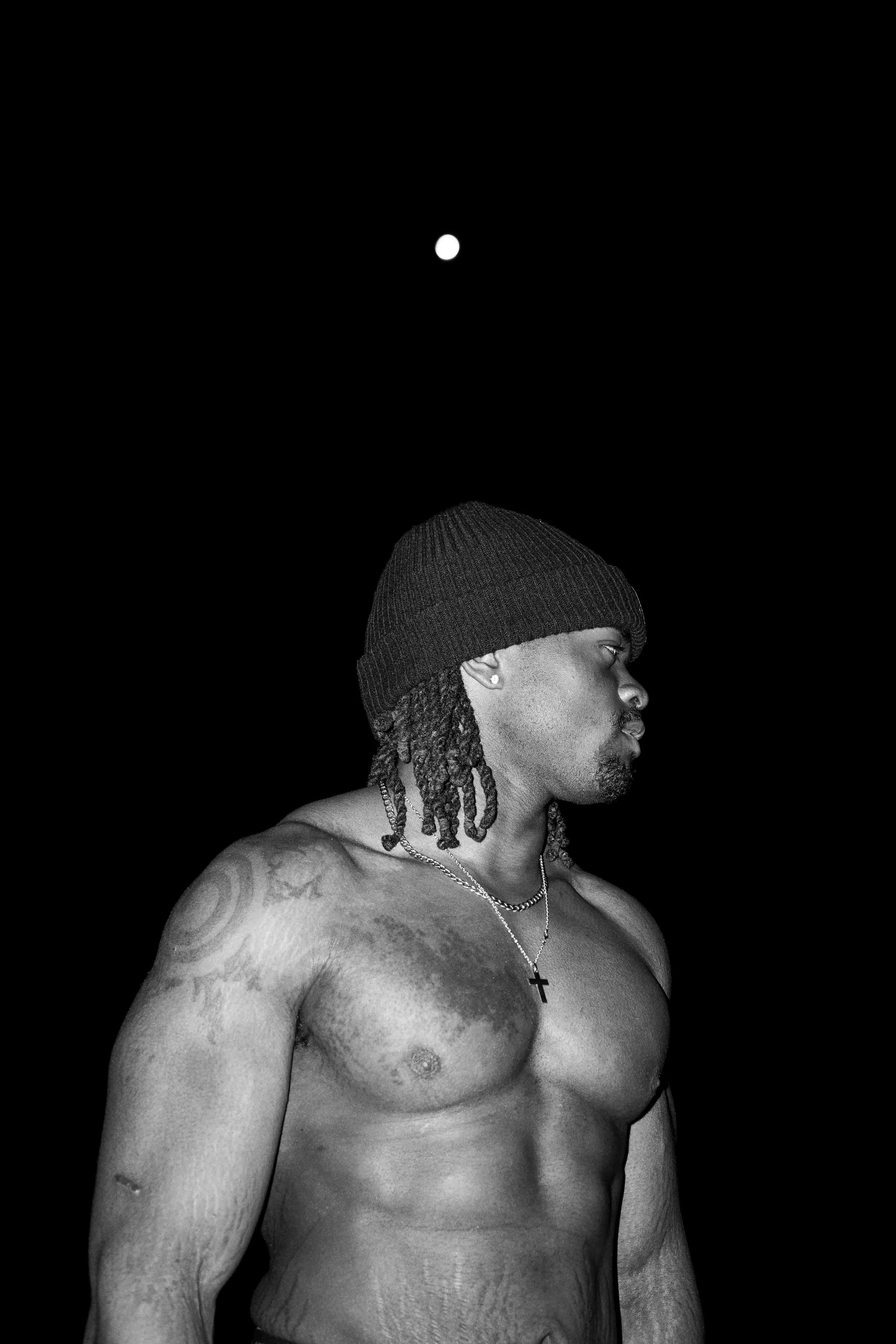 A shirtless man with tattoos, dreadlocks, wearing a black beanie and necklaces, looking to the side against a black background with a visible moon.