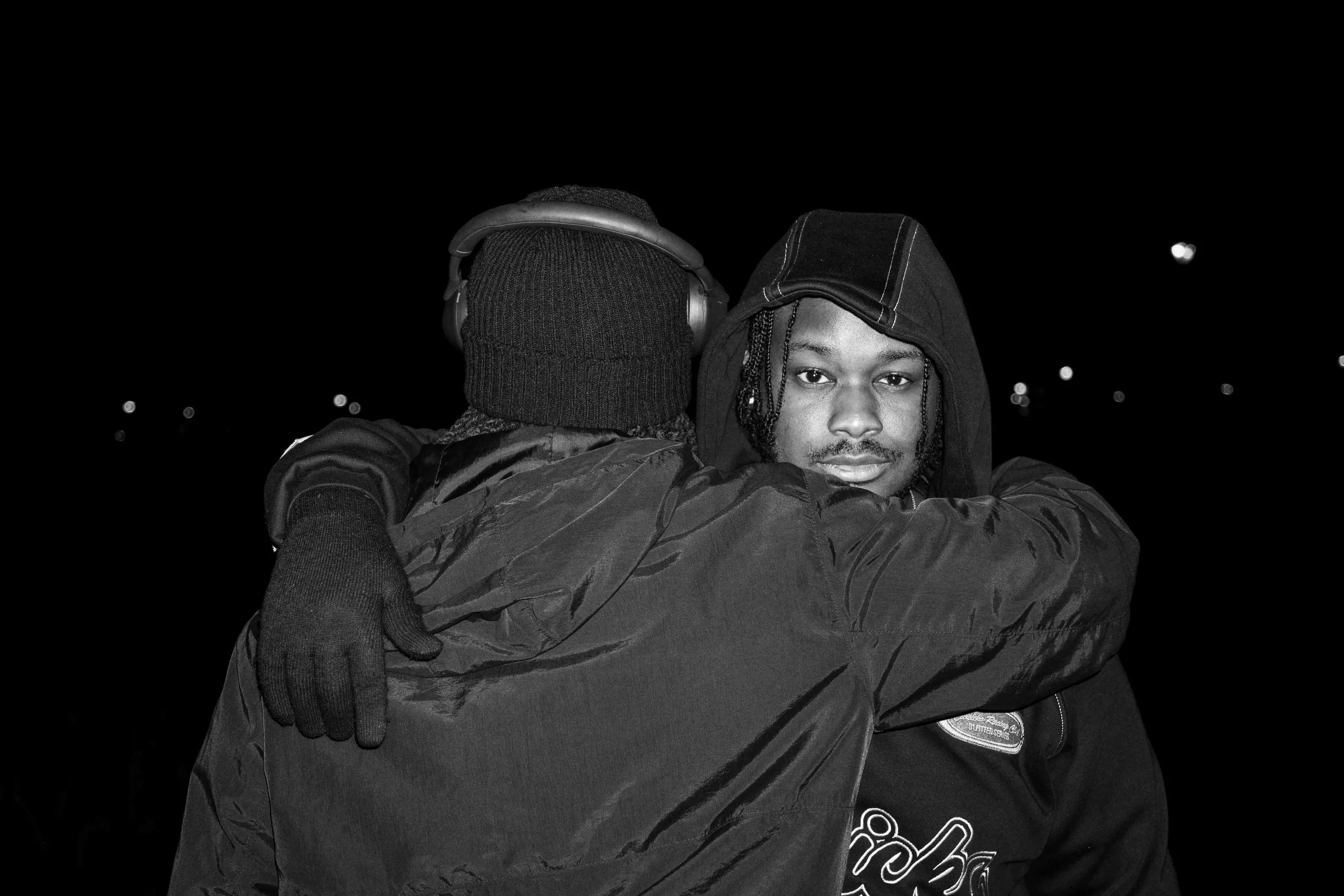 Two men embracing at night, one with a hoodie and the other with headphones and a knit cap, dark background with distant lights