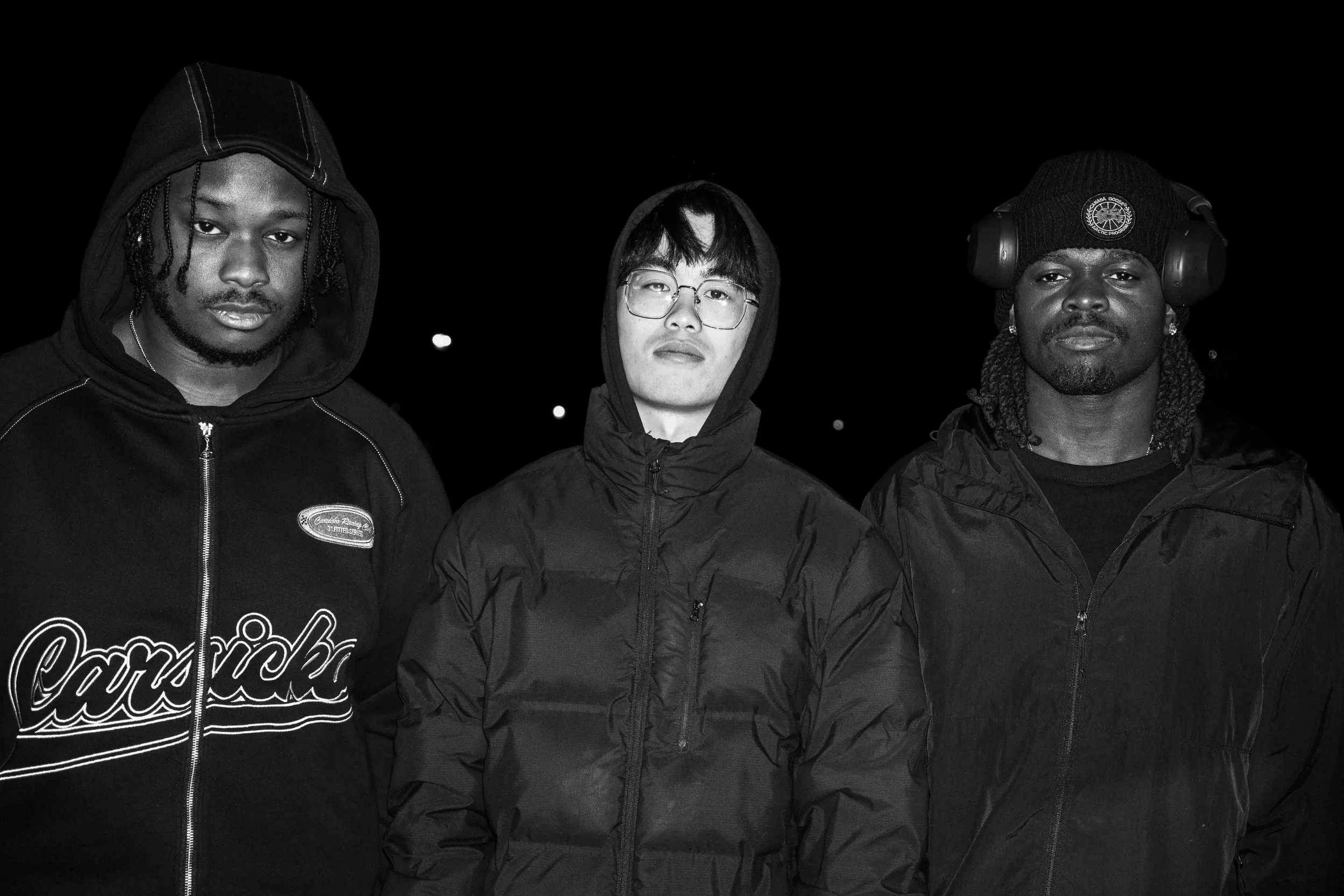 Three young men standing outdoors at night, dressed in dark hooded jackets with serious expressions.