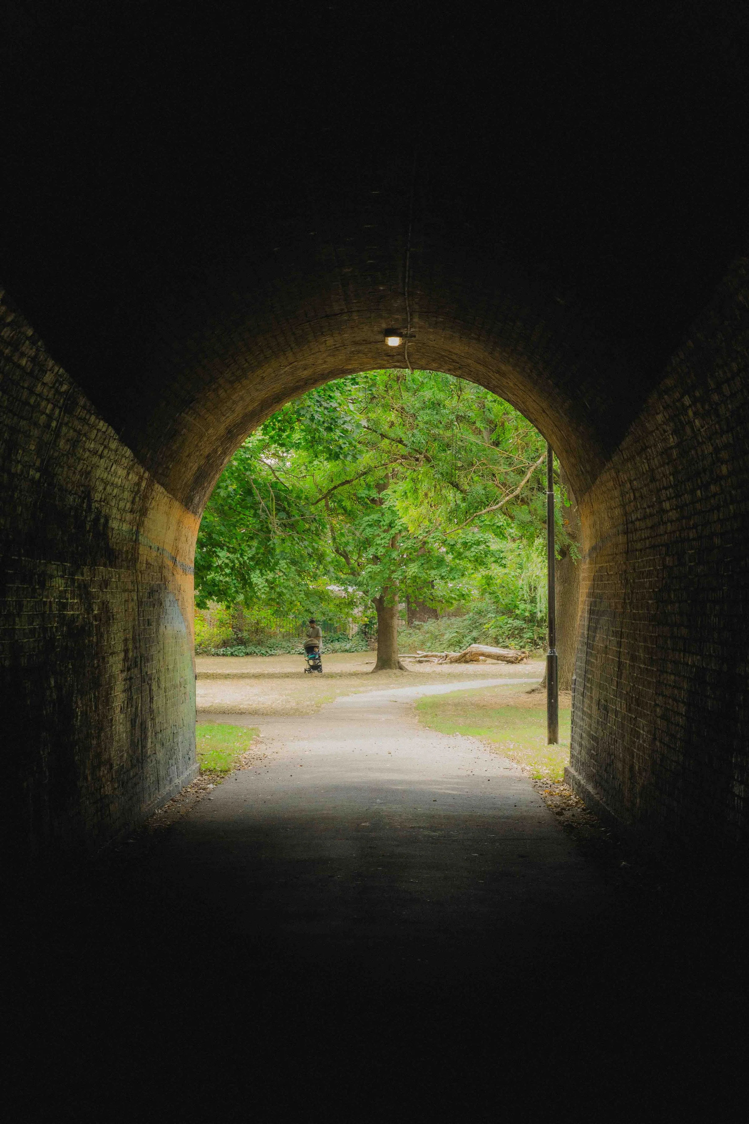 View from inside a dark tunnel looking out to a park with grass, trees, to a person sitting on a bench, and a person pushing a stroller.