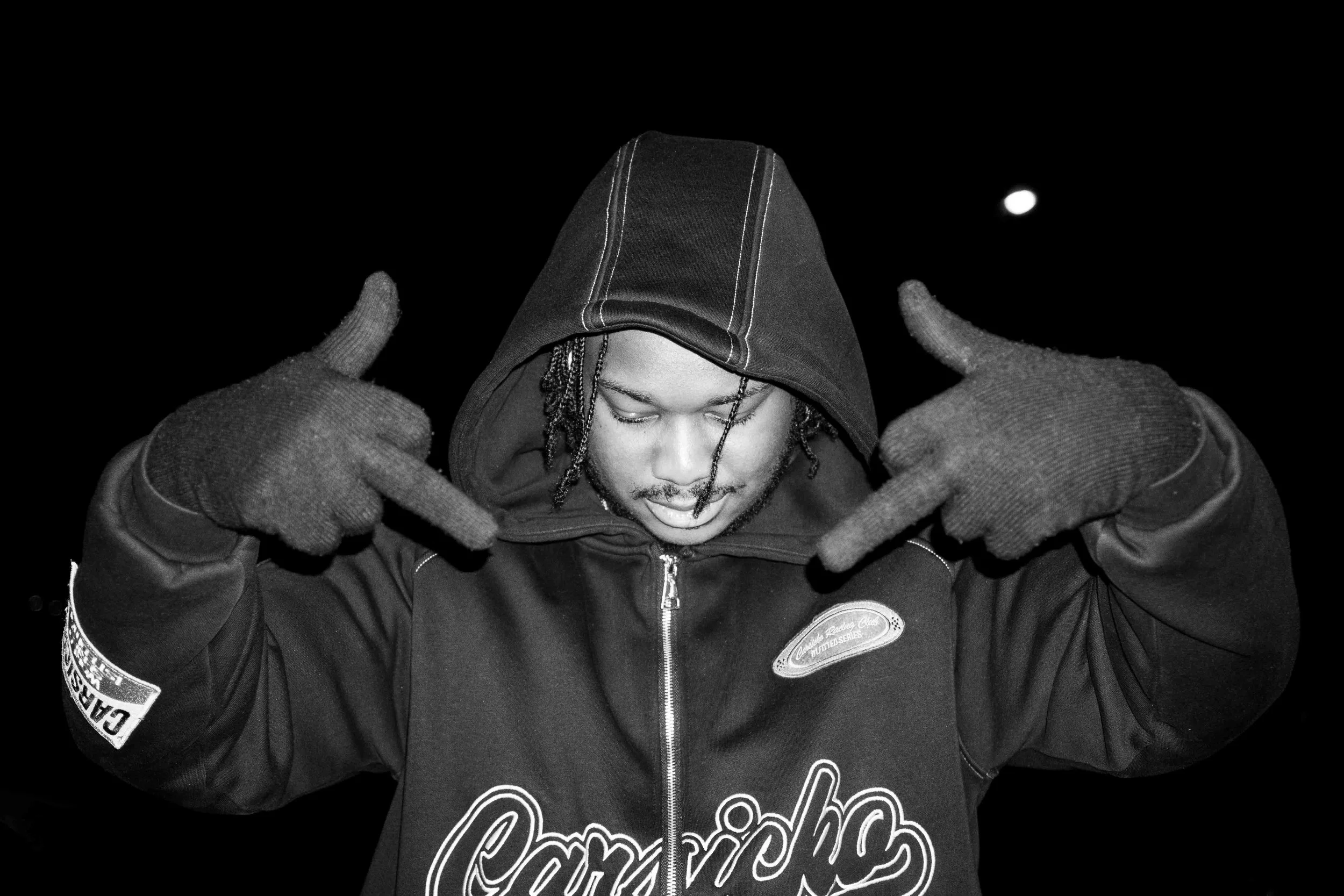 A young man wearing a hooded jacket and gloves making a hand gesture, with his head slightly bowed and eyes closed, against a dark background.