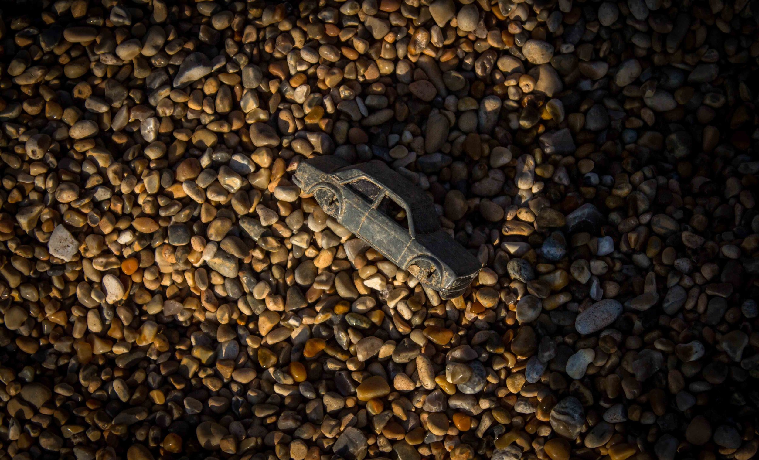 A small toy car lying on a bed of smooth, multicolored pebbles in natural sunlight.