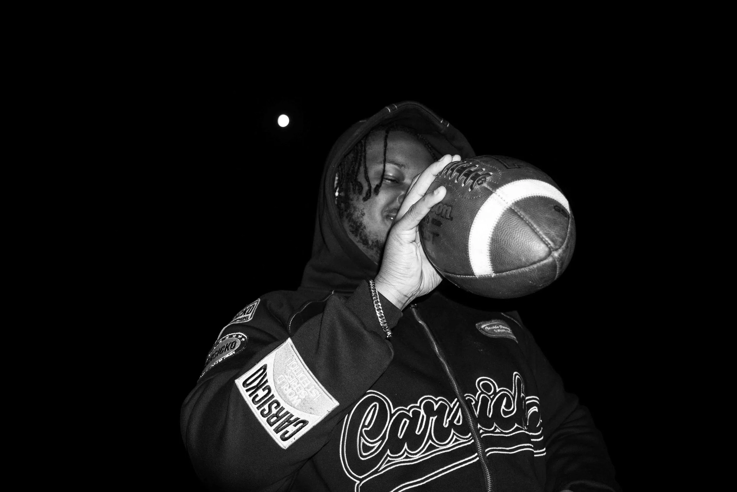 A young man in a baseball hoodie holding a football to his face, with the moon visible in the night sky behind him.