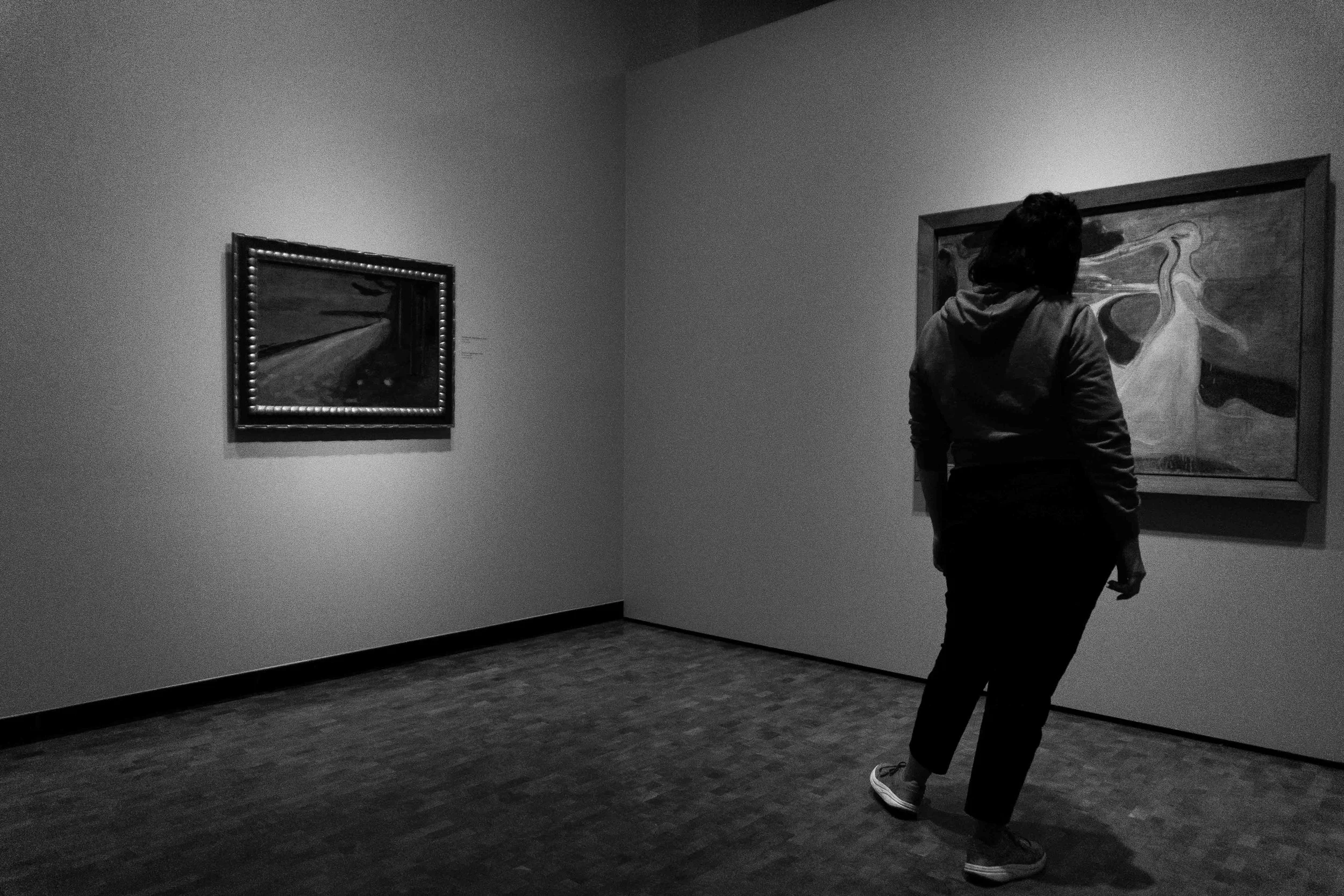 A person wearing a hoodie and sneakers standing in an art gallery, looking at a large painting on the wall.