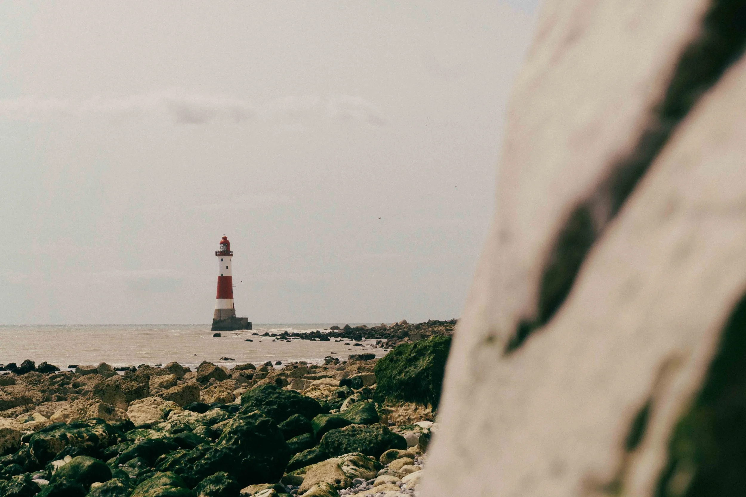 A lighthouse with red and white stripes standing in the ocean, seen from a rocky shoreline with a blurred white structure in the foreground.