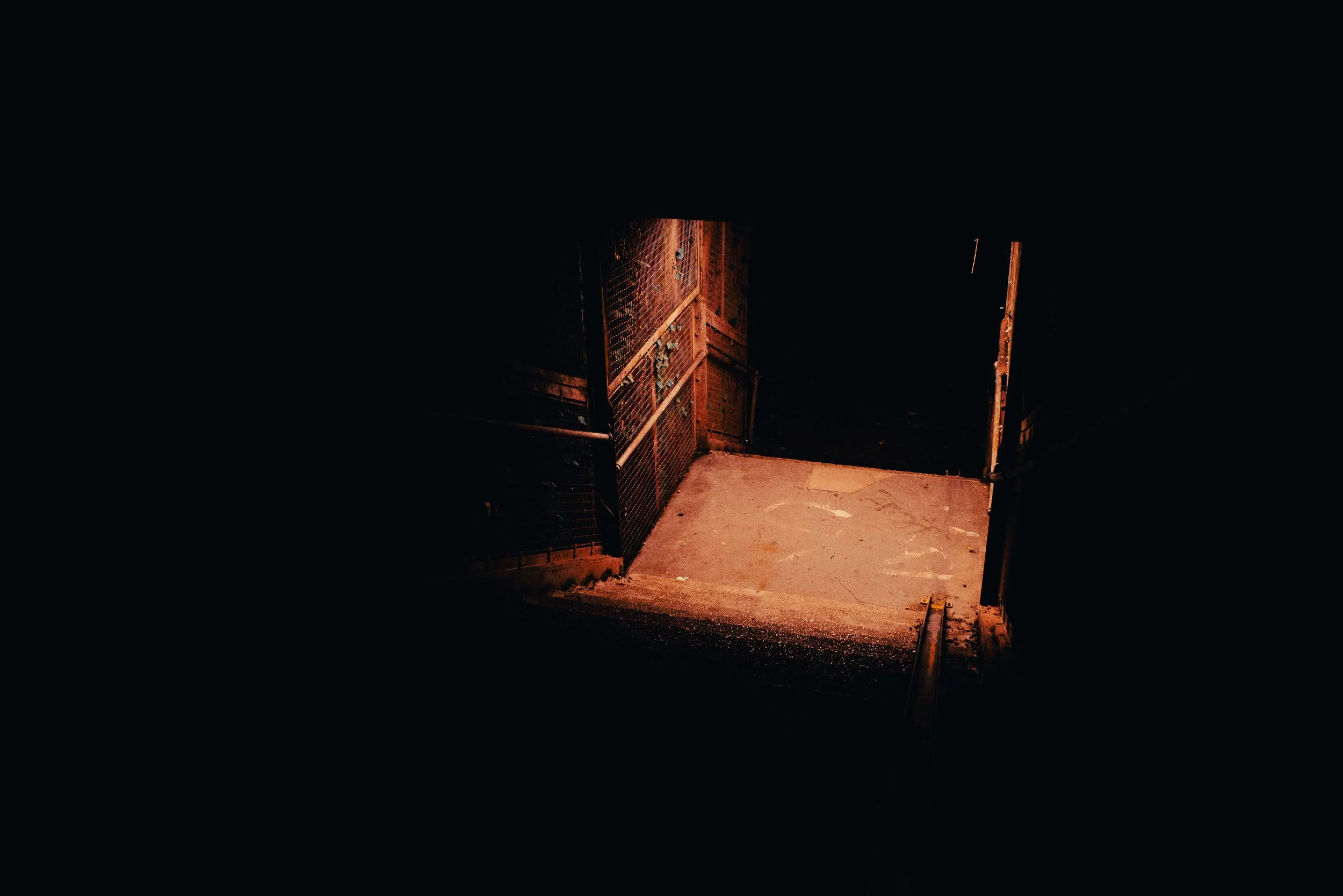 A dark stairway leading down to a small landing with dim, reddish lighting, metal railings, and concrete steps.