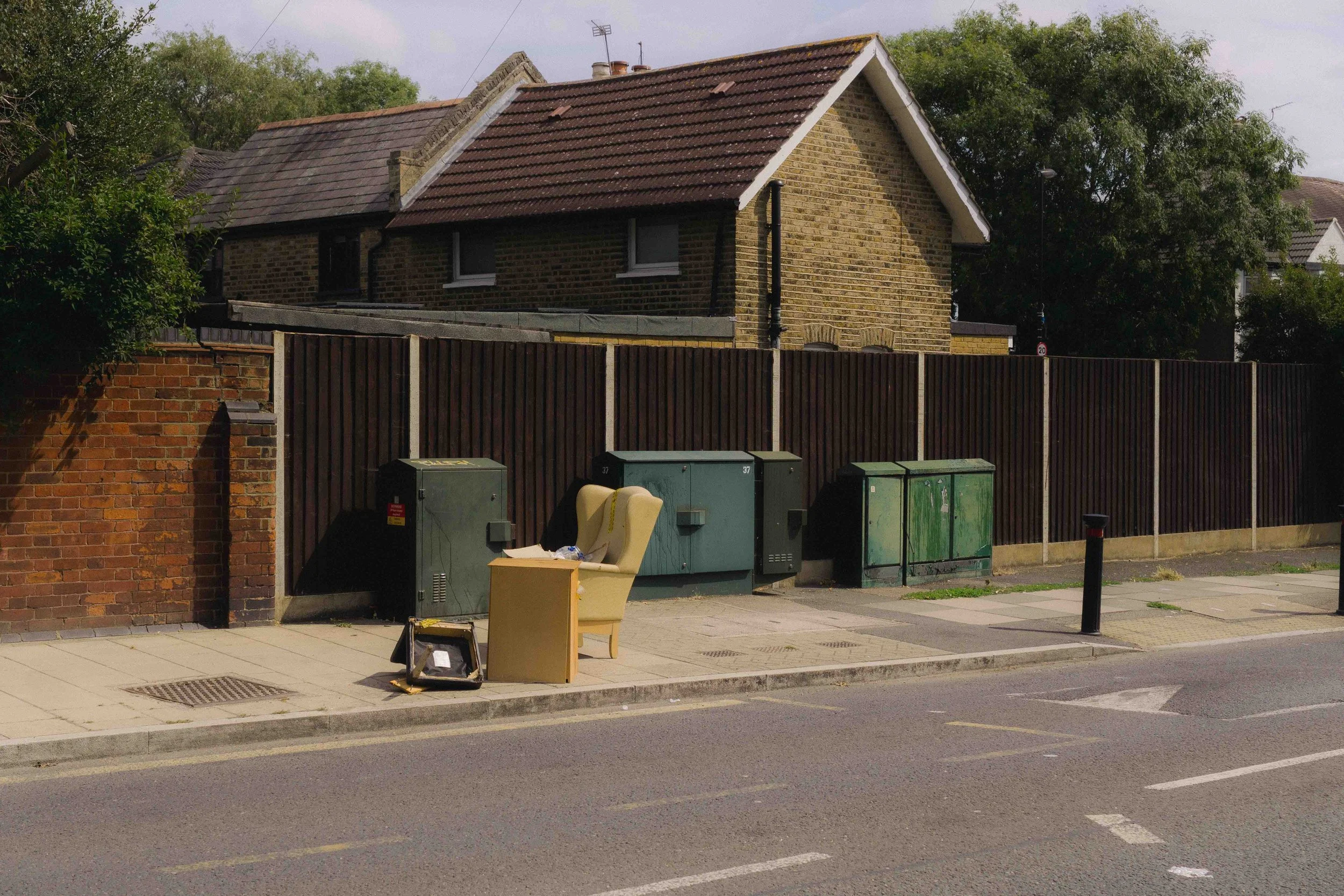 A sidewalk with abandoned furniture, including a chair, a desk, and a bag, with electrical boxes and a wooden fence behind. Residential houses and trees are visible in the background.
