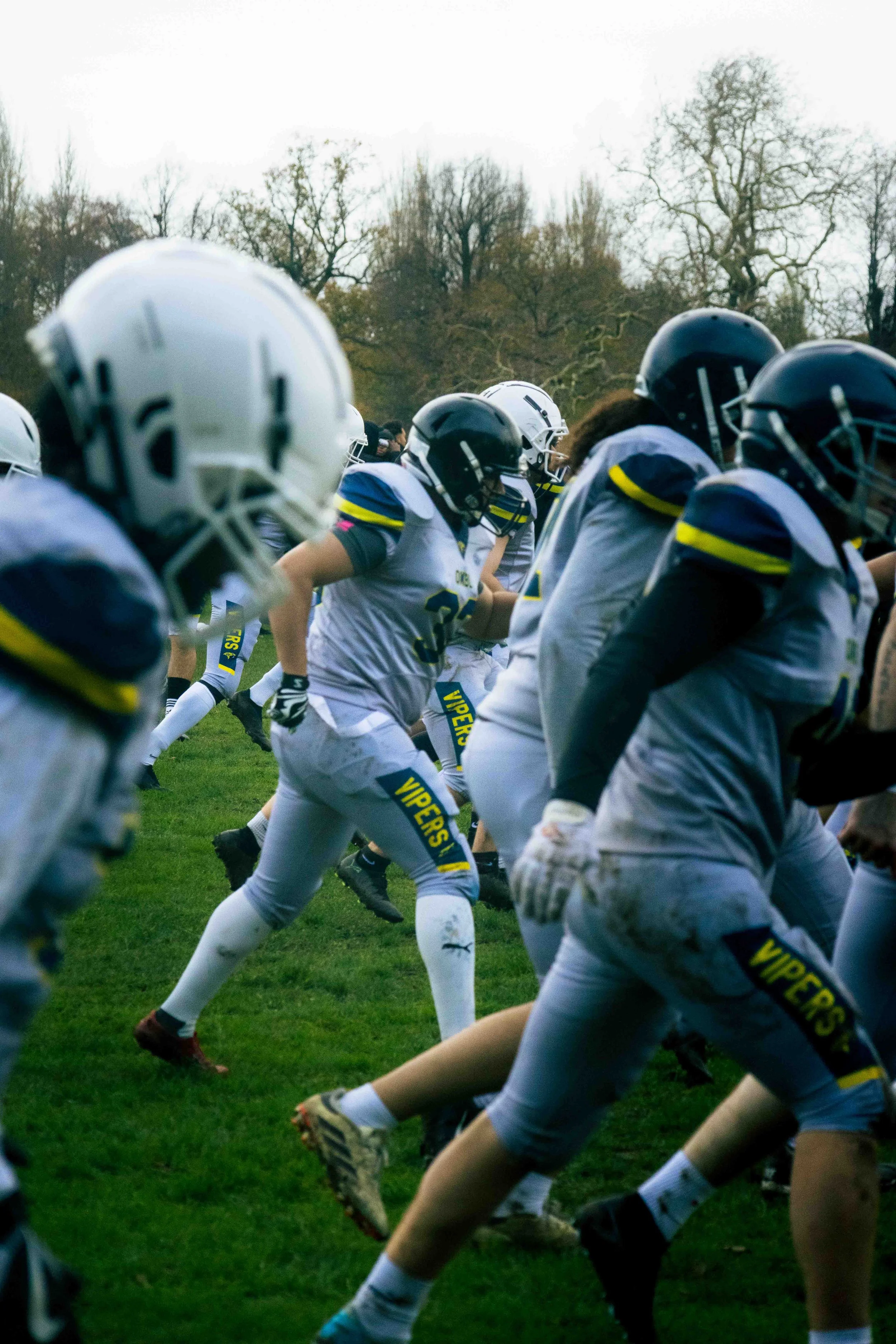 A group of football players, dressed in white and blue uniforms with helmets, are running onto the field during a game or practice. The players are in motion with trees in the background.