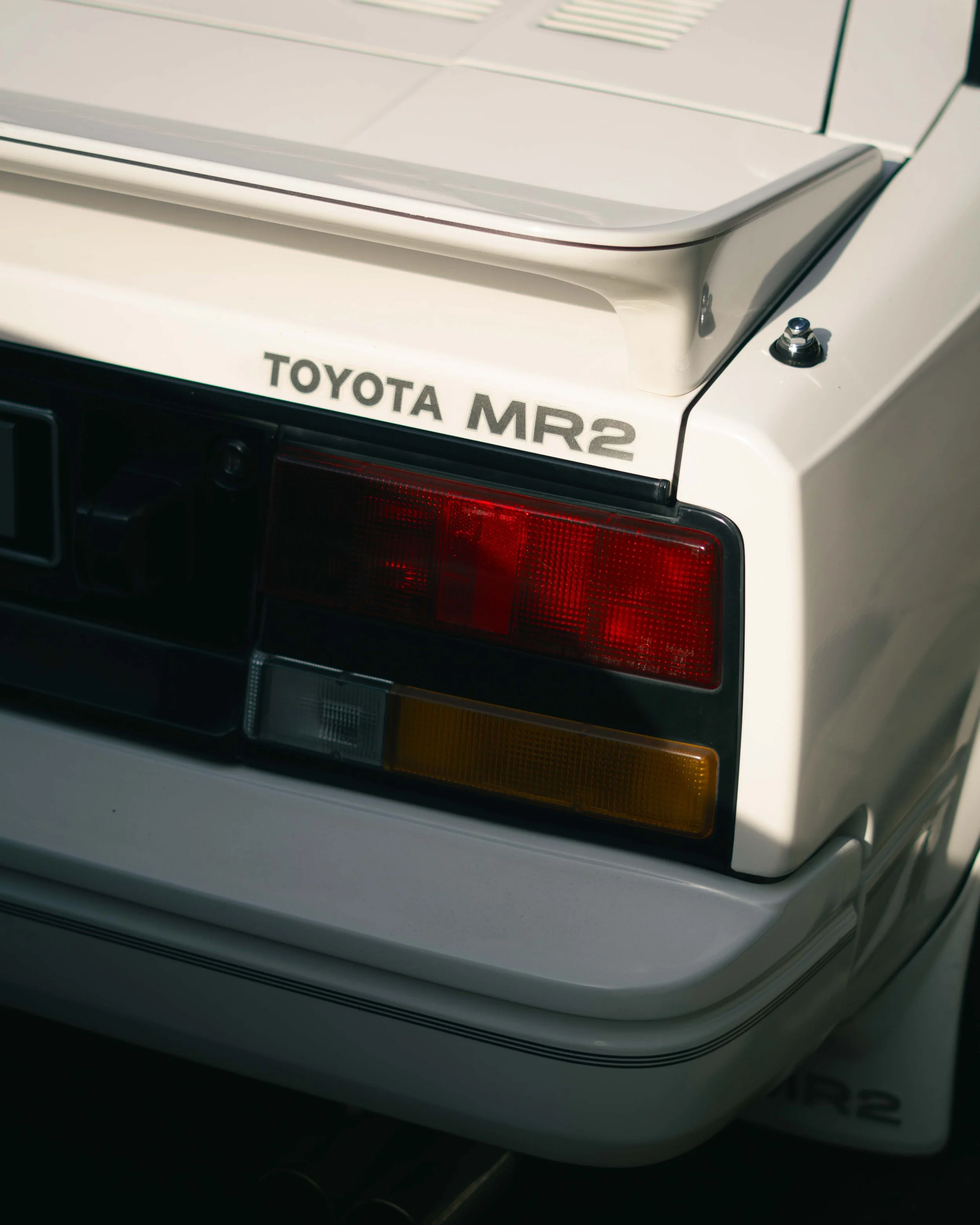 Close-up of the rear corner of a white Toyota MR2 car showing the tail light, black and white body, and the 'TOYOTA MR2' badge.