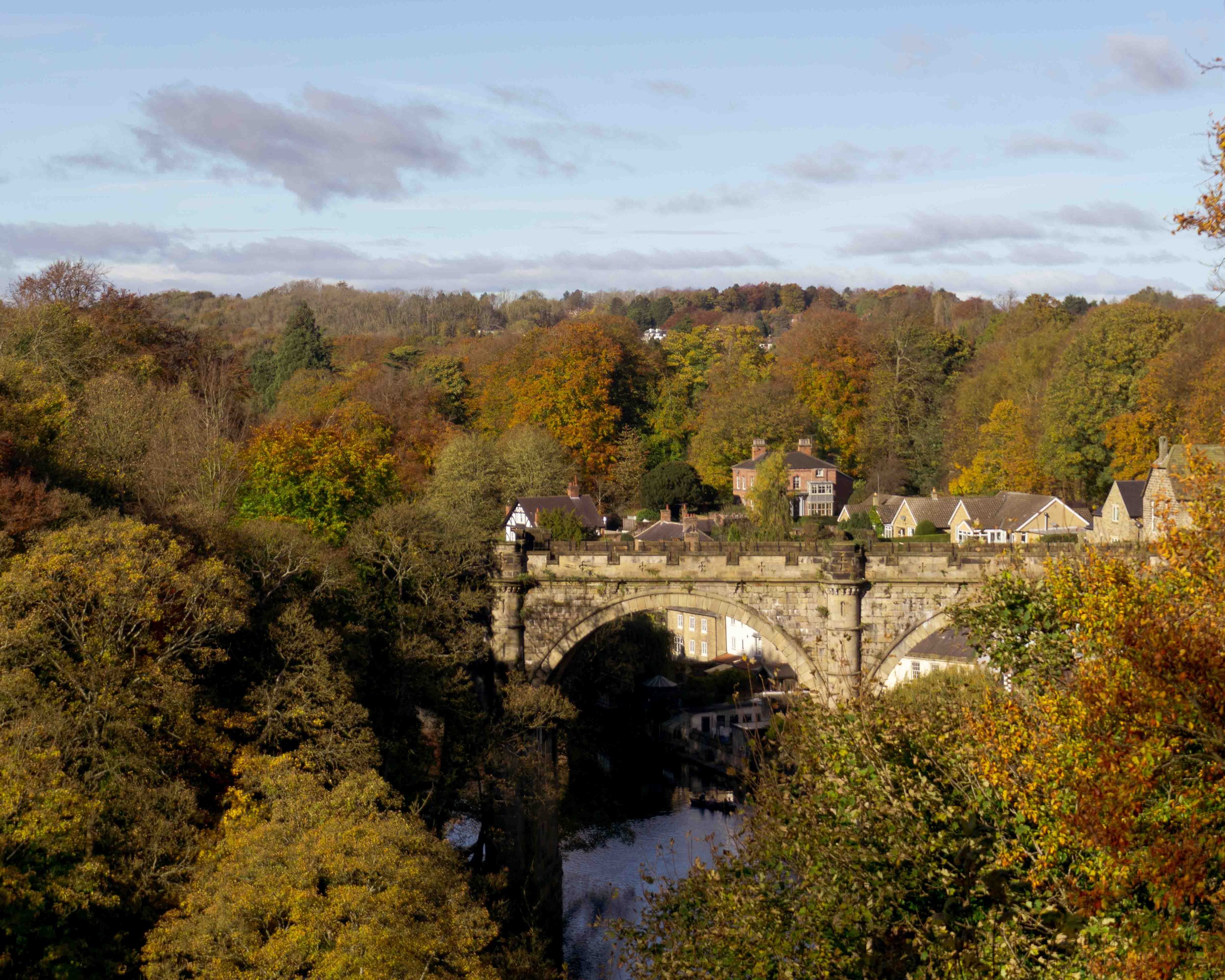 A scenic view of a historic stone bridge over a river, surrounded by colorful trees and houses with autumn foliage.