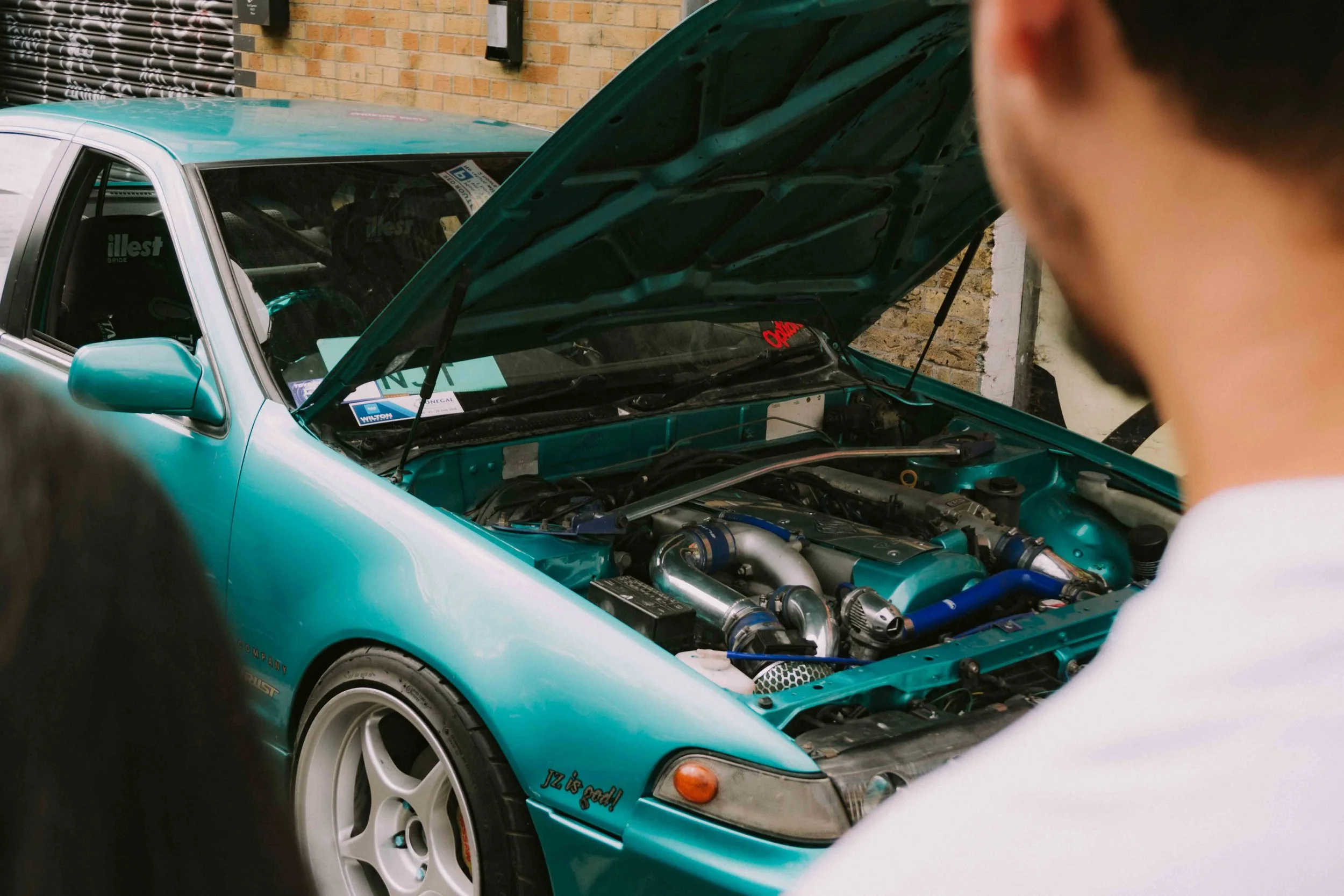 A teal sports car with its hood open, exposing a modified engine, parked near a brick wall, with people observing.