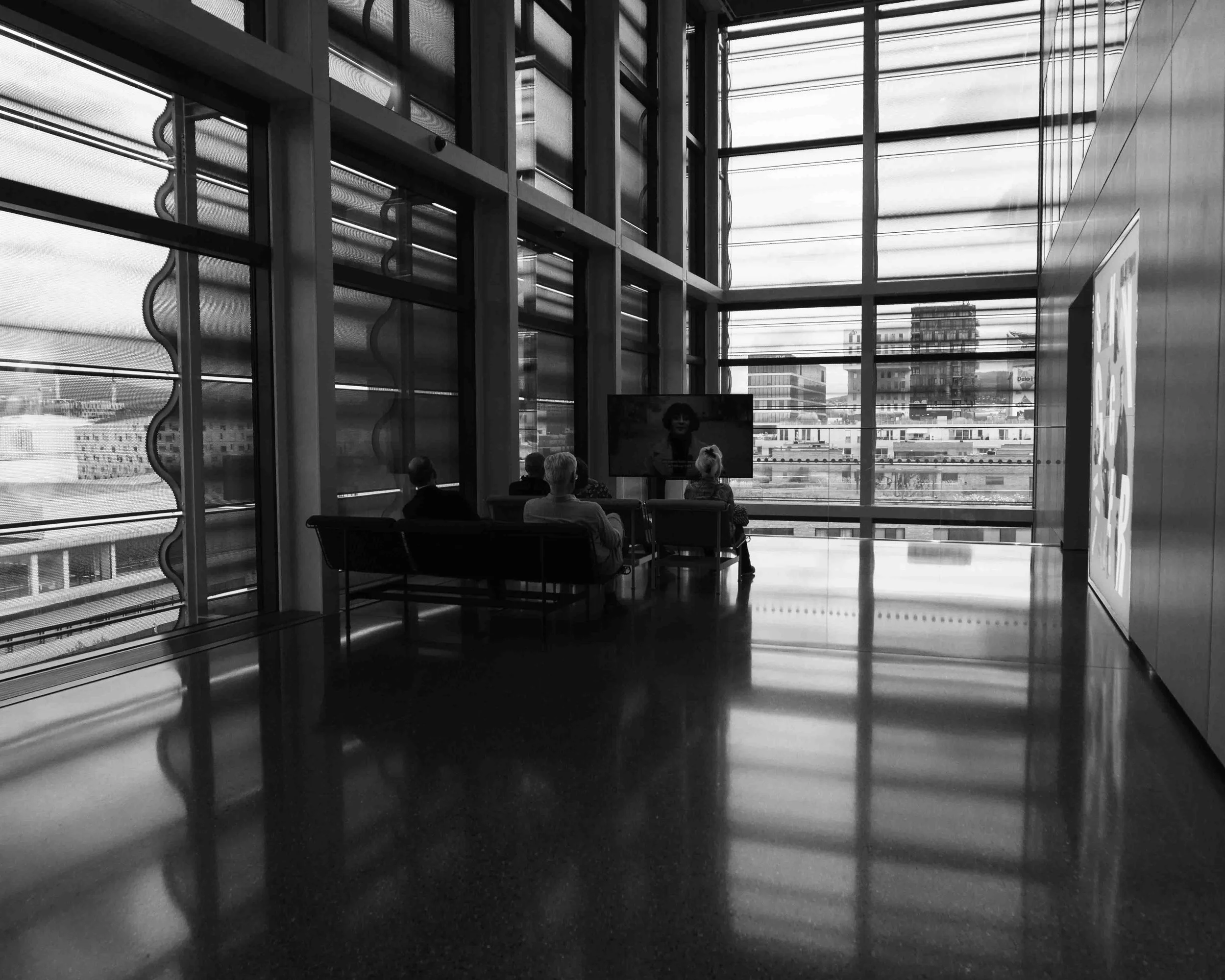 People sitting in chairs watching a television in a modern building with large glass windows, cityscape outside.