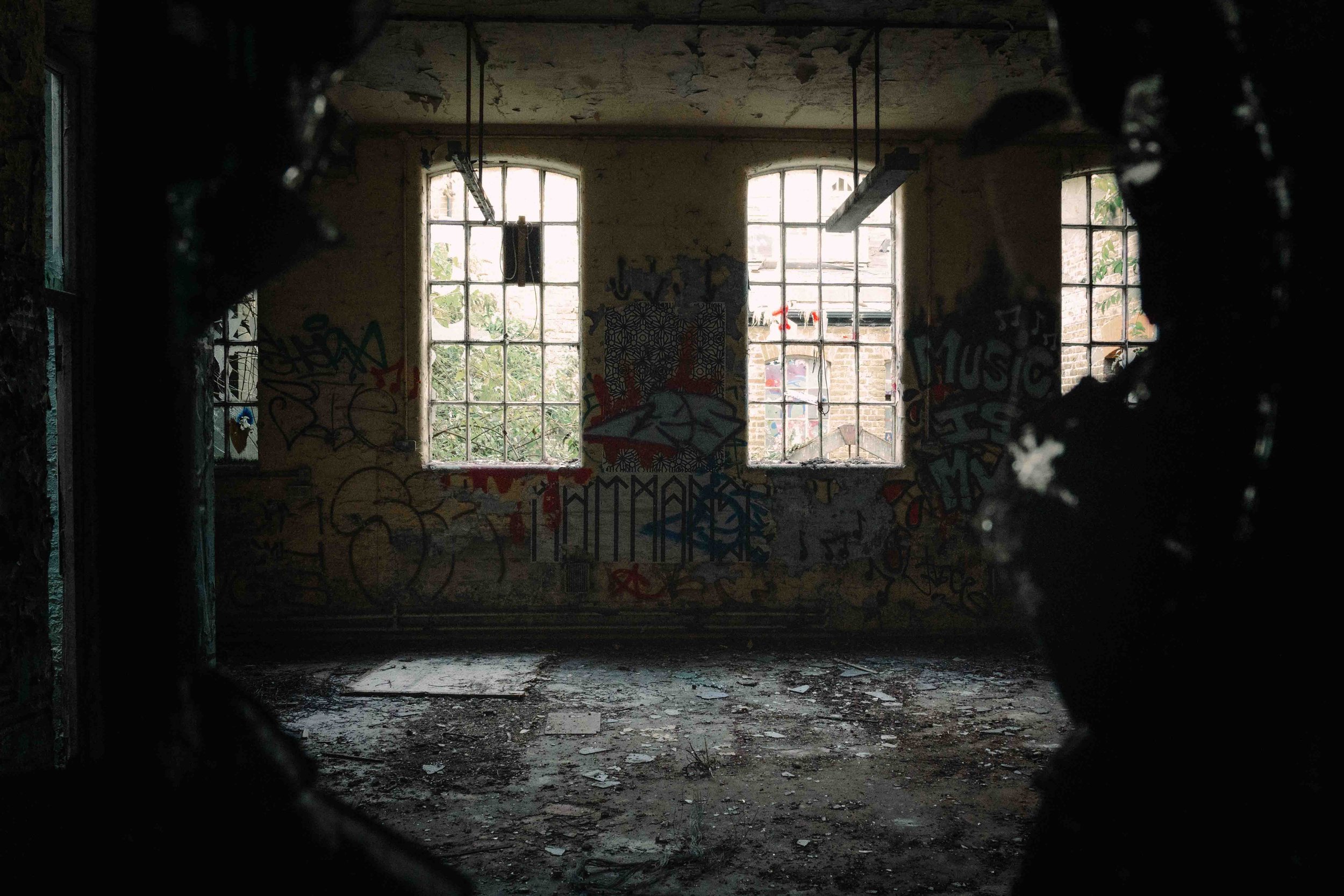Interior of an abandoned building with three large windows, graffiti on the walls, and debris on the floor, seen through a broken opening.
